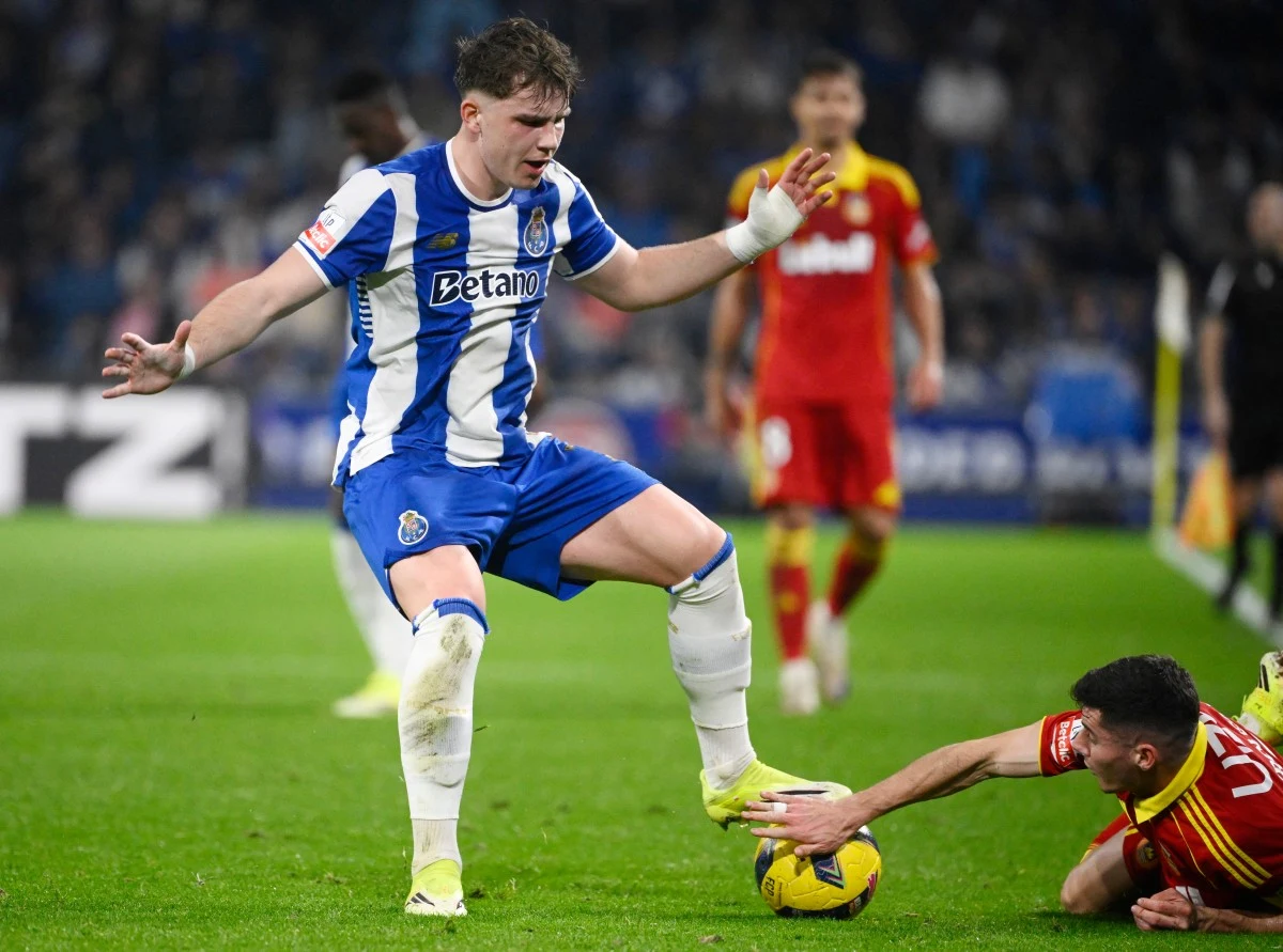 Zawodnik drużyny w biało-niebieskim stroju próbuje utrzymać się przy piłce, podczas gdy zawodnik przeciwnej drużyny w czerwono-żółtym stroju leży na murawie, sięgając ręką po piłkę, a w tle stoją inni piłkarze.