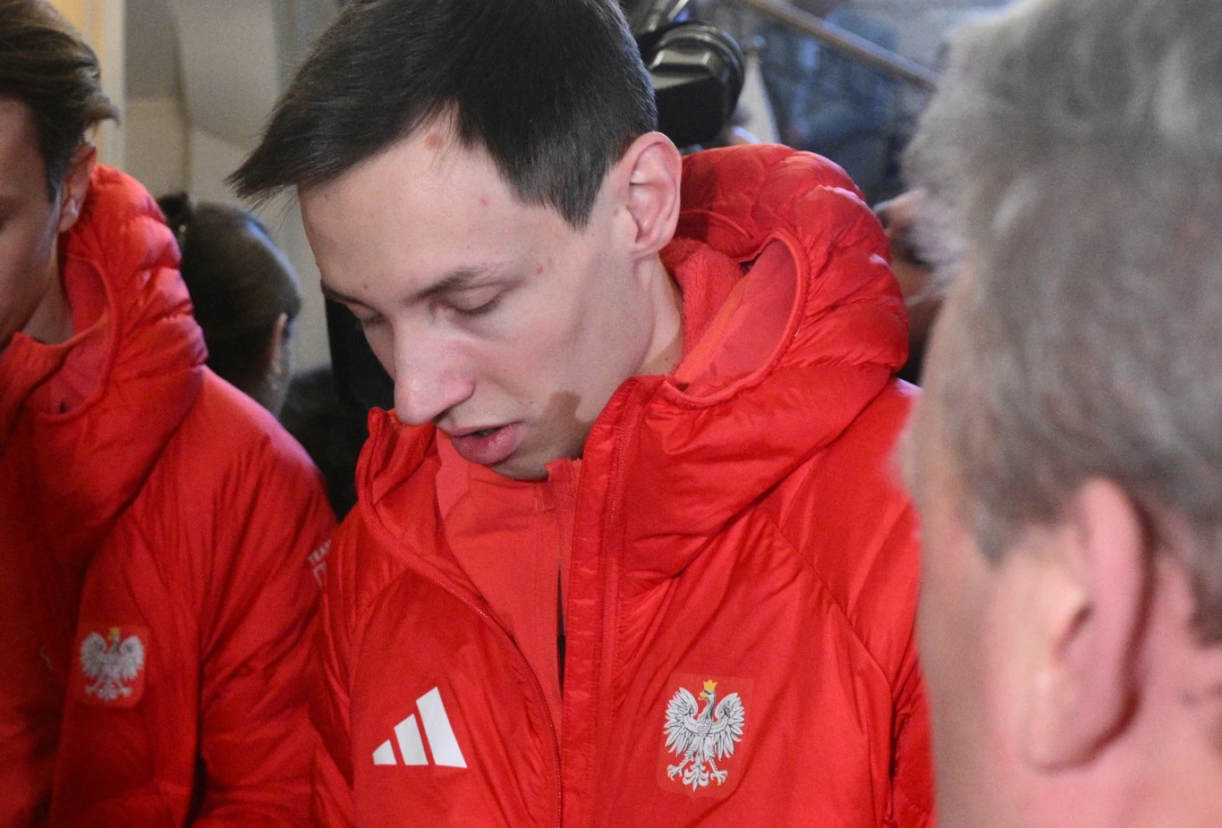 Kacper Tomasiak Młody mężczyzna ubrany w czerwoną kurtkę z godłem Polski oraz logo sportowej marki, pochylony do przodu, otoczony przez inne osoby w podobnych kurtkach.