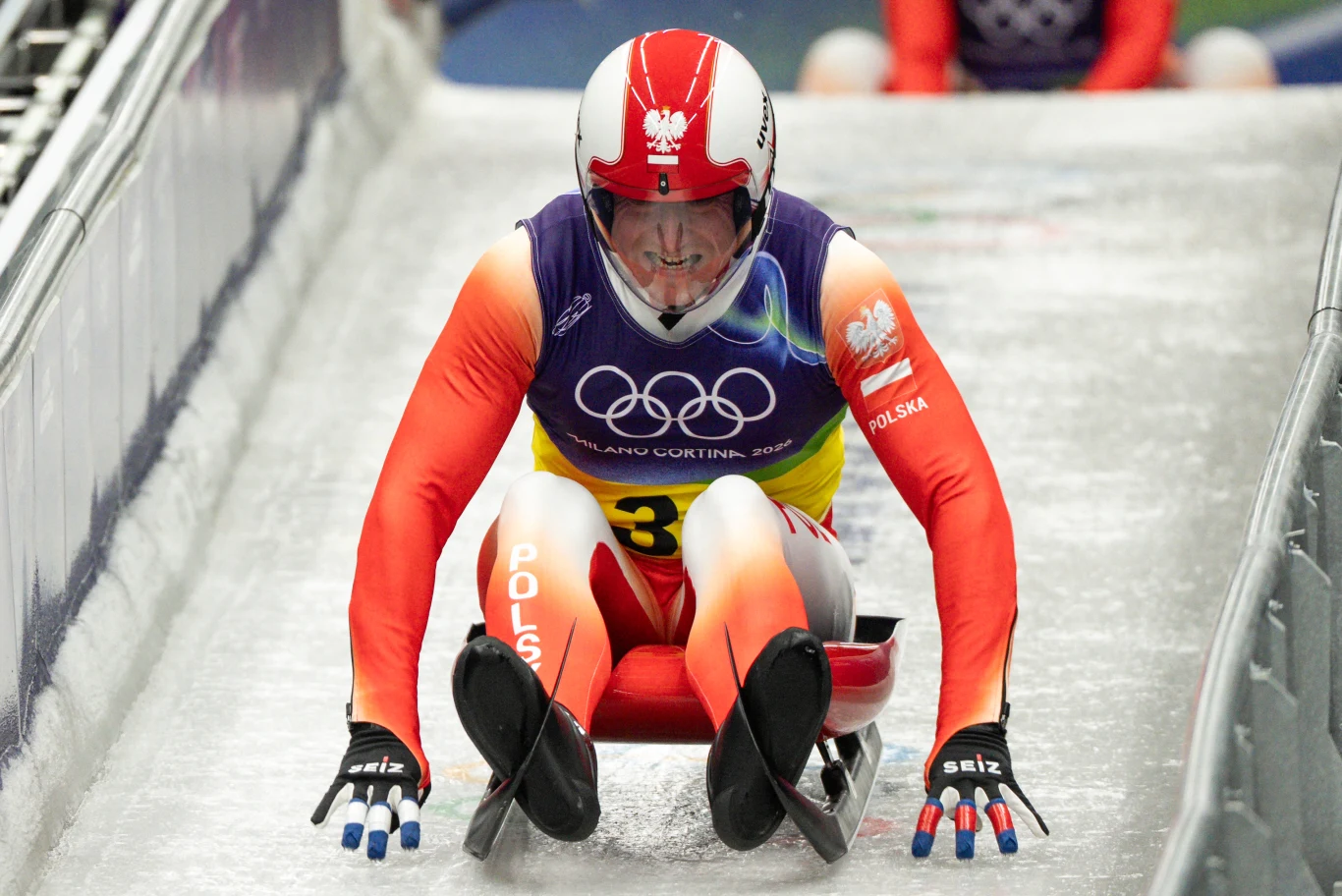 Zjazd saneczkarski zawodnika z Polski w specjalnym stroju i kasku na torze lodowym podczas zawodów olimpijskich, charakterystyczne logo igrzysk widoczne na tle stroju.