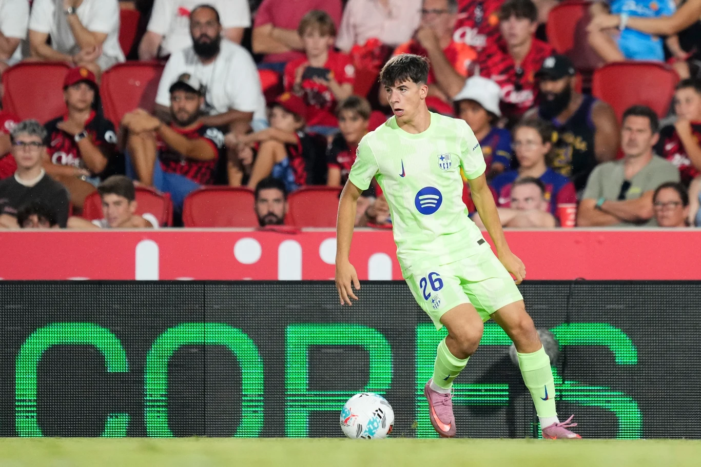 Piłkarz w jasnożółtym stroju kontroluje piłkę na tle trybun pełnych kibiców podczas meczu piłkarskiego na stadionie.