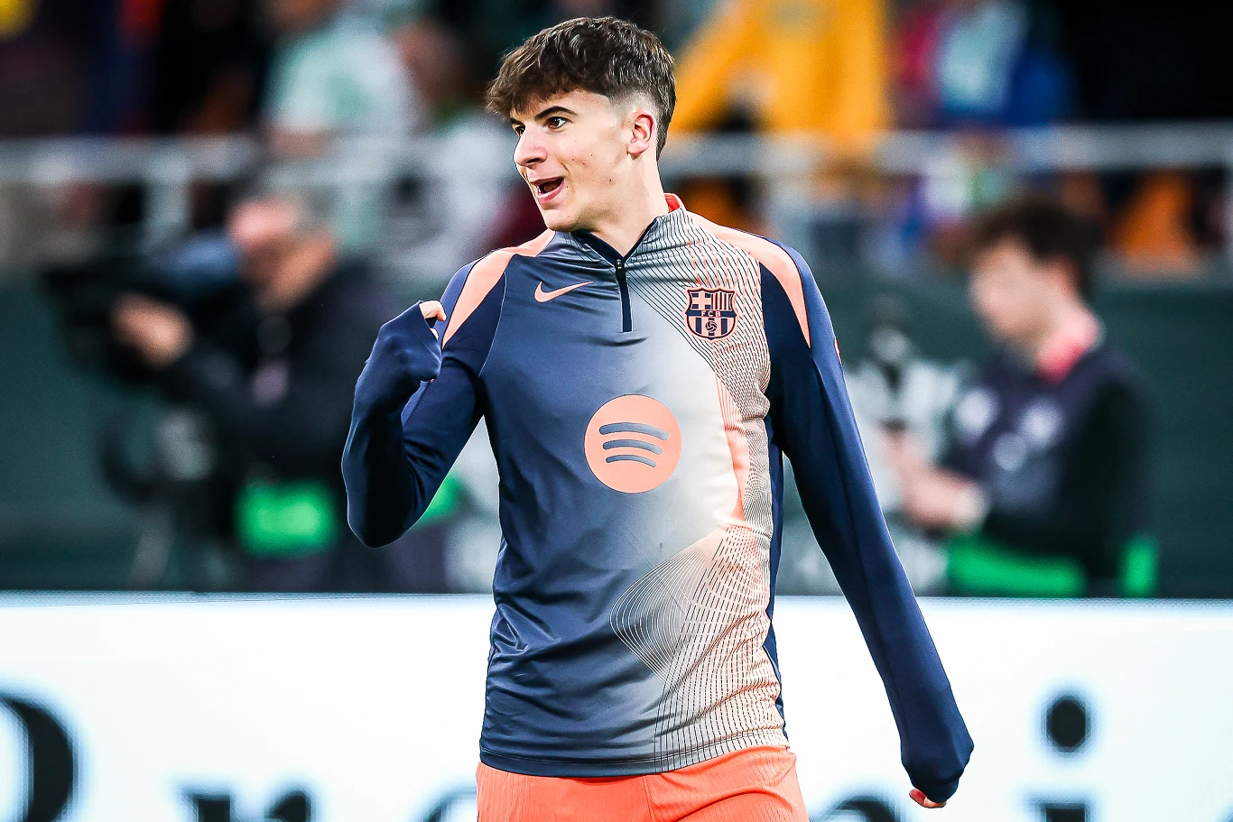 Młody mężczyzna w sportowym stroju FC Barcelony z widocznym logo sponsora Spotify, stoi na tle niewyraźnej, stadionowej publiczności, unosi rękę i uśmiecha się.
