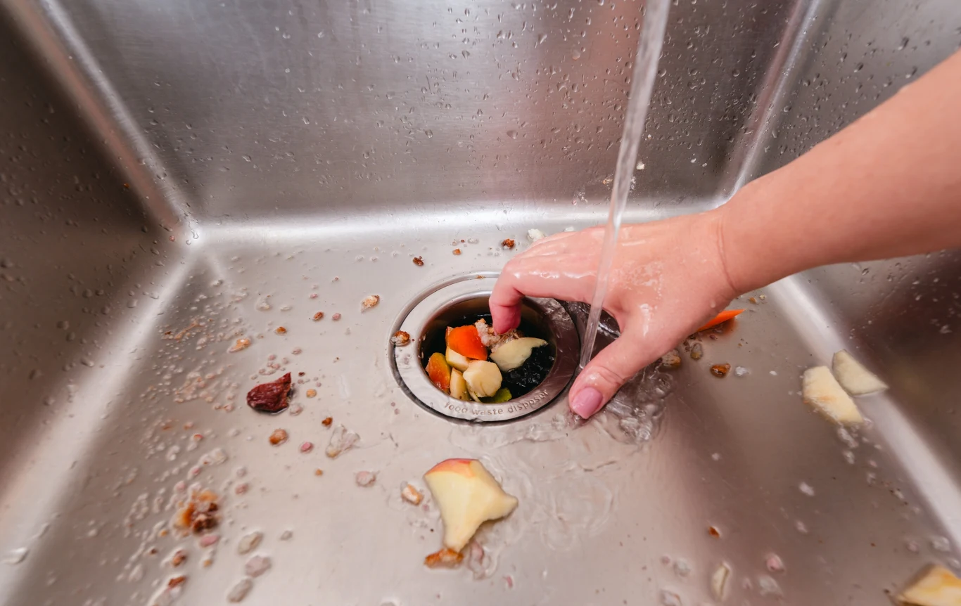 "Kanalizacja nie jest miejscem na odpady kuchenne" - wyjaśnia pracowniczka oczyszczalni Ręka osoby zbierająca resztki jedzenia wokół otworu odpływowego w zlewie kuchennym, przez który płynie woda, wokół widoczne kawałki obierek i resztek jedzenia.