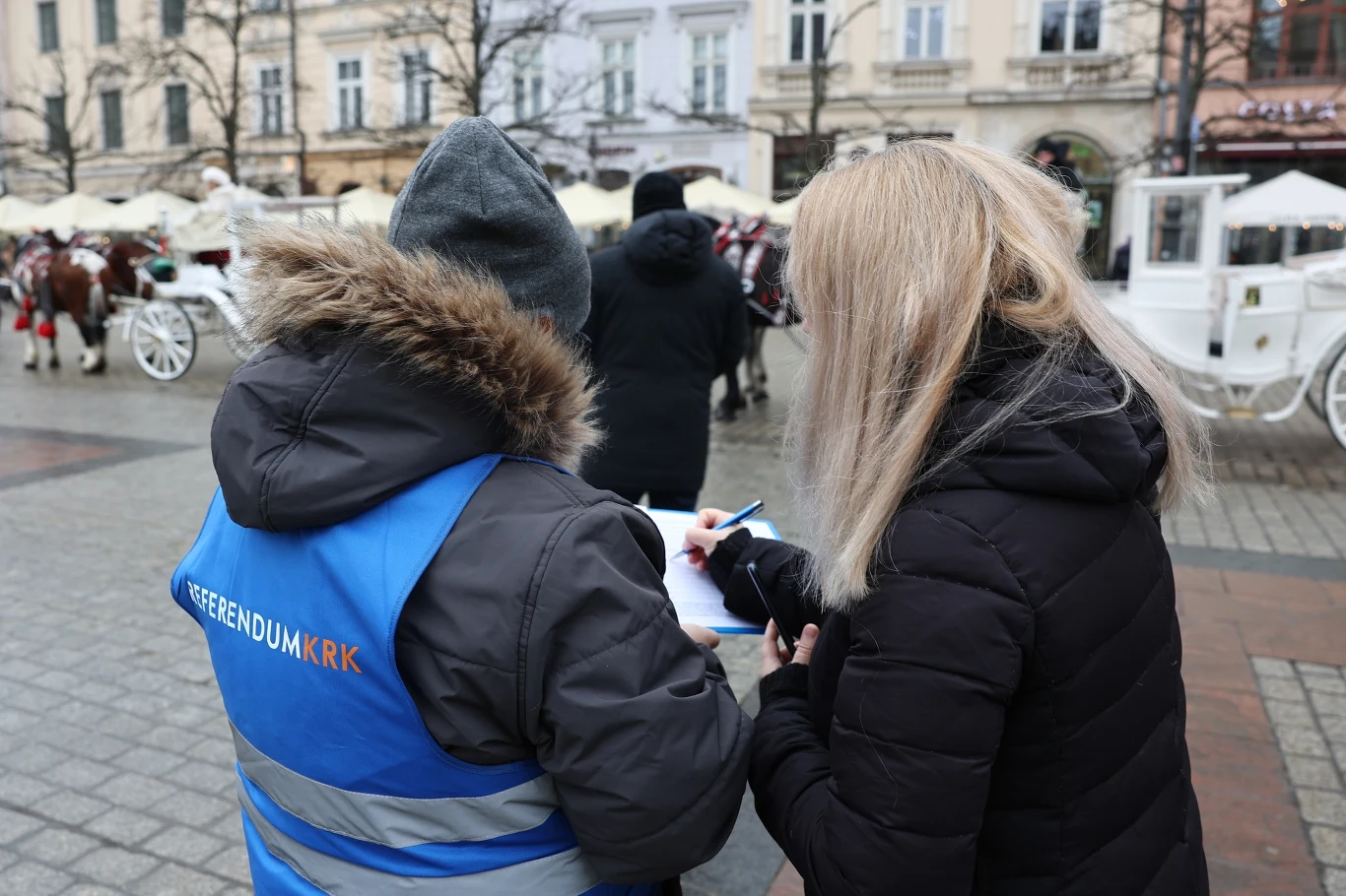 Dwie osoby stoją na rynku miejskim, jedna z nich ubrana w niebieską kamizelkę z napisem Referendum KRK, obie pochylają się nad dokumentem i coś zapisują, w tle jasne kamienice oraz białe dorożki zaprzężone w konie.