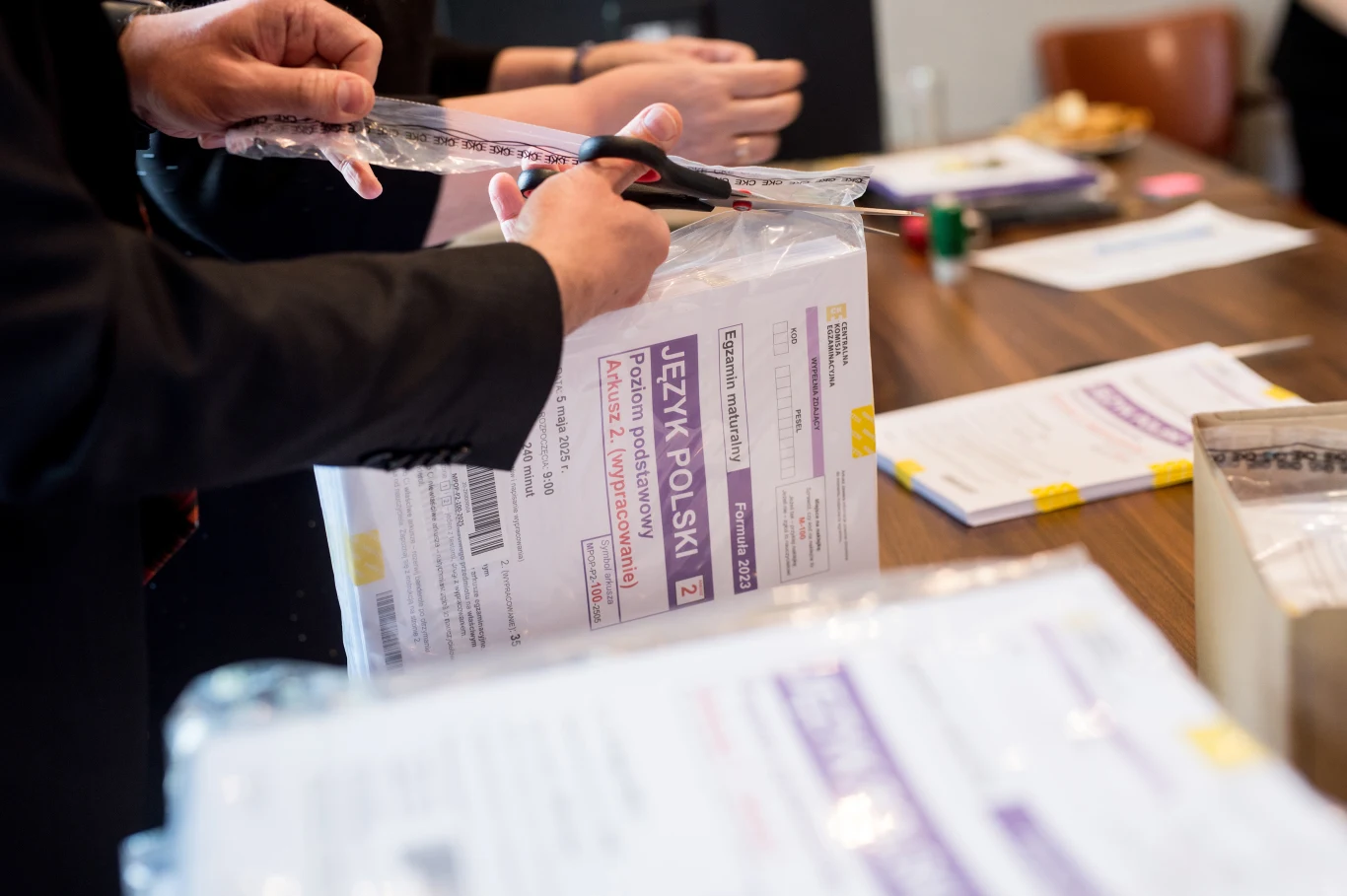 Osoba otwiera pakiet egzaminacyjny z języka polskiego za pomocą nożyczek, na stole leżą kolejne arkusze. 