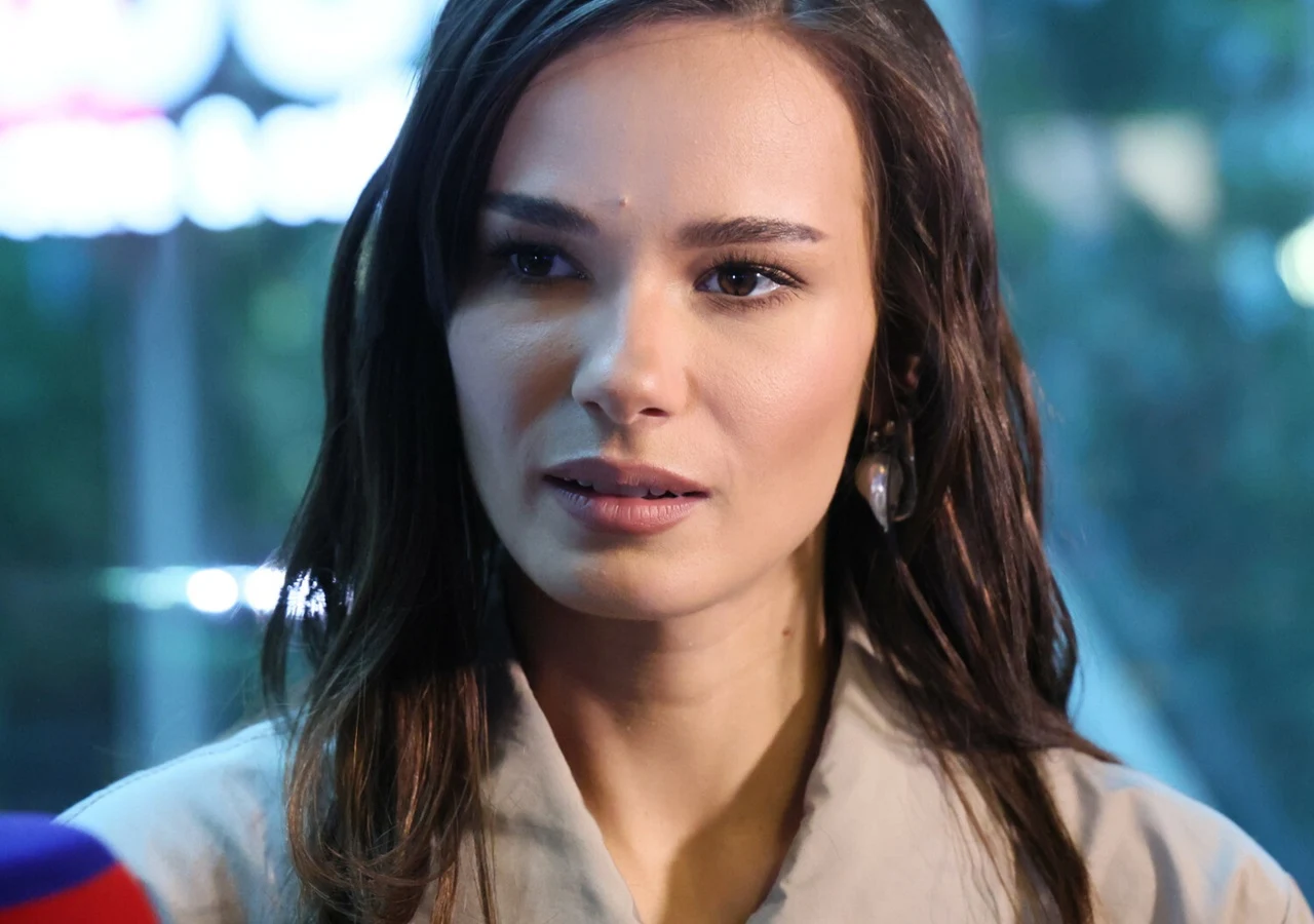 Natalia Szroeder Młoda kobieta o długich, ciemnych włosach patrzy lekko w bok, ubrana w jasną marynarkę, tło rozmyte z jasnymi światłami.