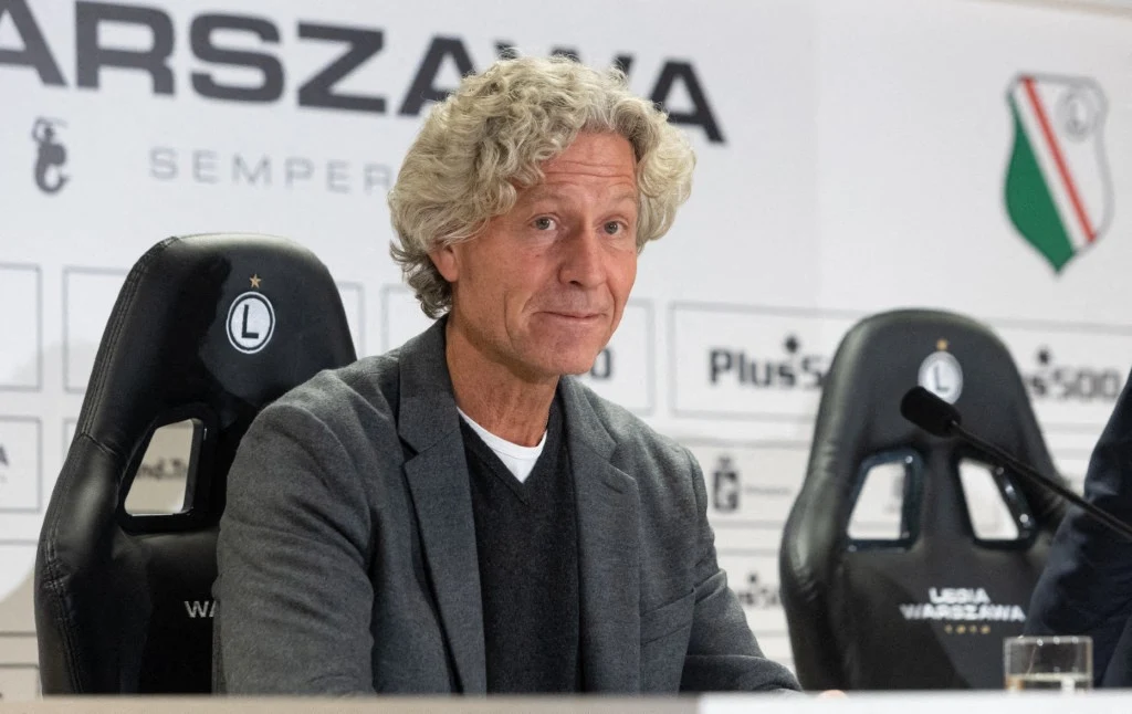 Mężczyzna z siwymi kręconymi włosami siedzi przy stole konferencyjnym na tle loga klubu piłkarskiego Legia Warszawa, ubran w szarą marynarkę i sweter.