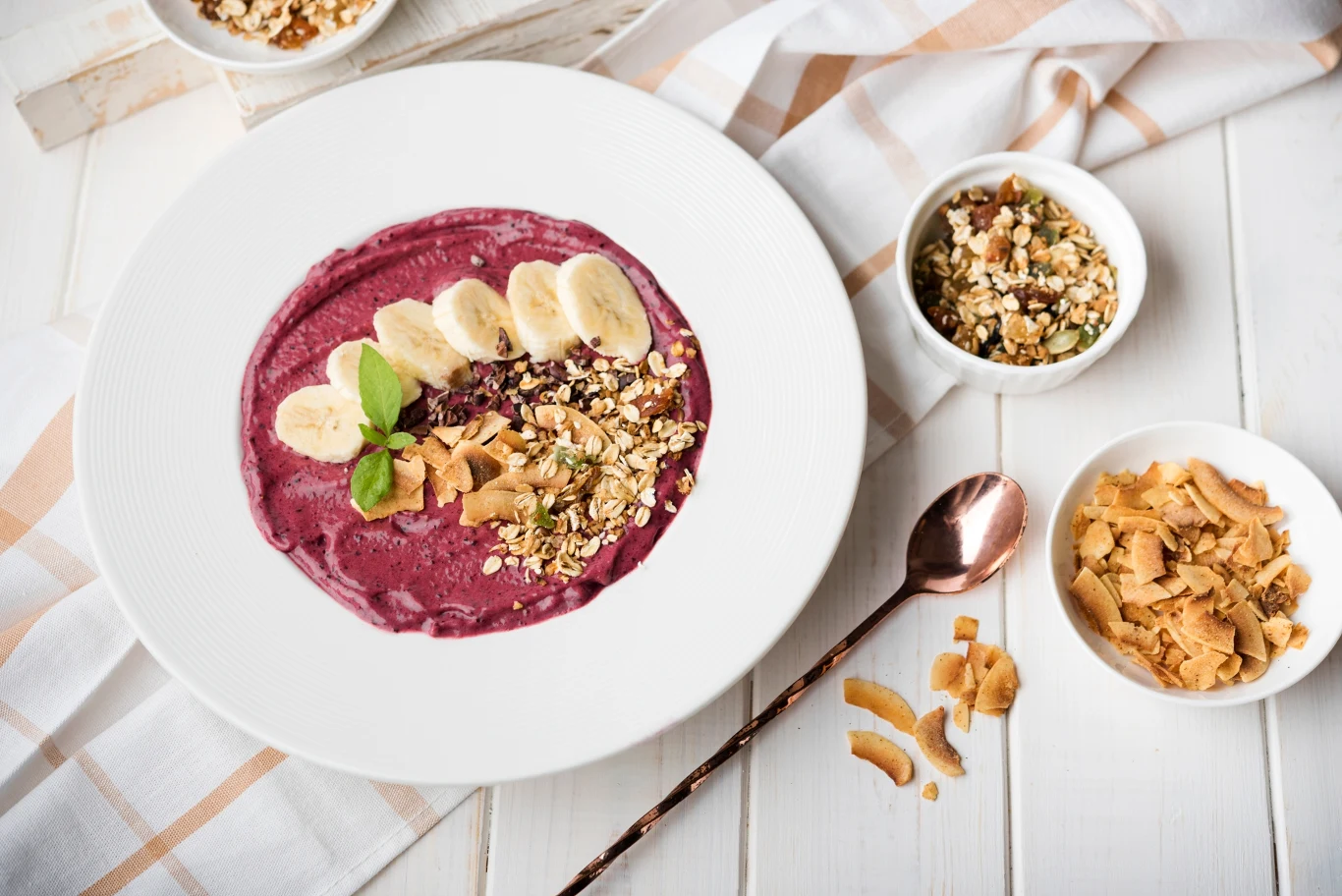 Smoothie bowl, czyli misa pełna zdrowia Smoothie-bowl jagodowe udekorowane plasterkami banana, płatkami kokosa, orzechami oraz świeżą bazylią. Obok miski znajdują się małe miseczki z dodatkami oraz łyżka na białym stole.
