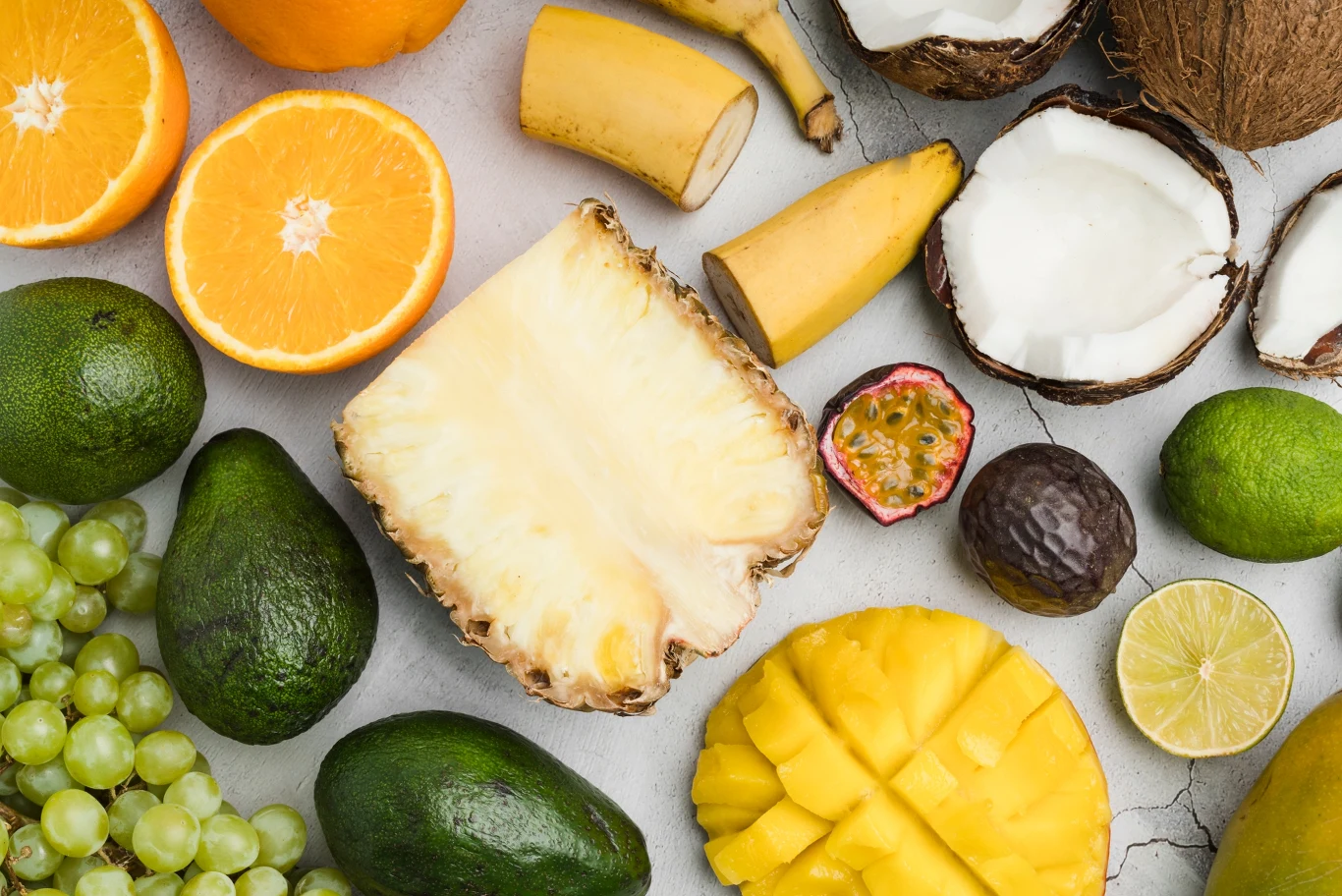 Pomysły na smoothie bowl Różnorodne egzotyczne owoce, takie jak ananas, awokado, mango, kokos, winogrona, pomarańcza, banan, limonka i marakuja, rozmieszczone na jasnym tle, większość z nich przekrojona na pół, ukazując świeże miąższe i nasiona.