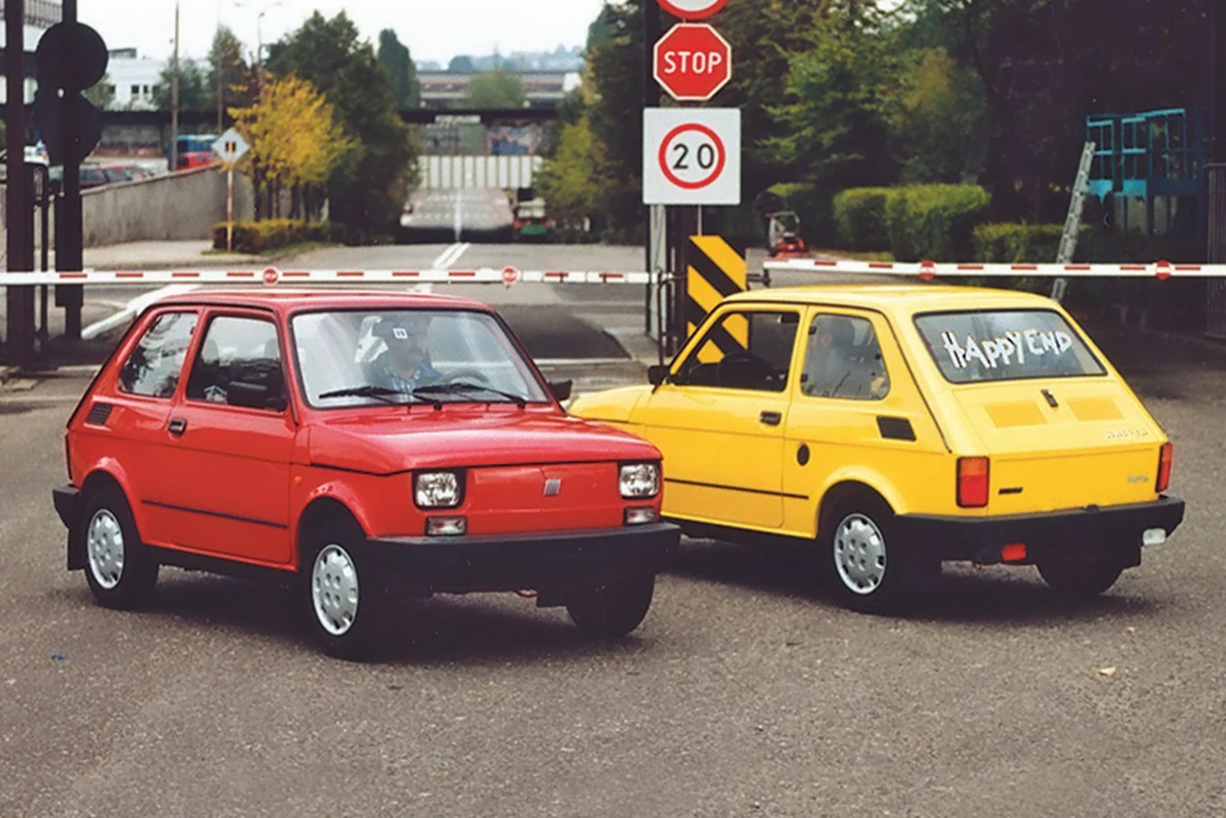 Dwa małe samochody Fiat 126p, jeden czerwony, drugi żółty, ustawione na asfaltowej nawierzchni przed rogatką; na szybie tylnej żółtego auta napis 'Happy End'.