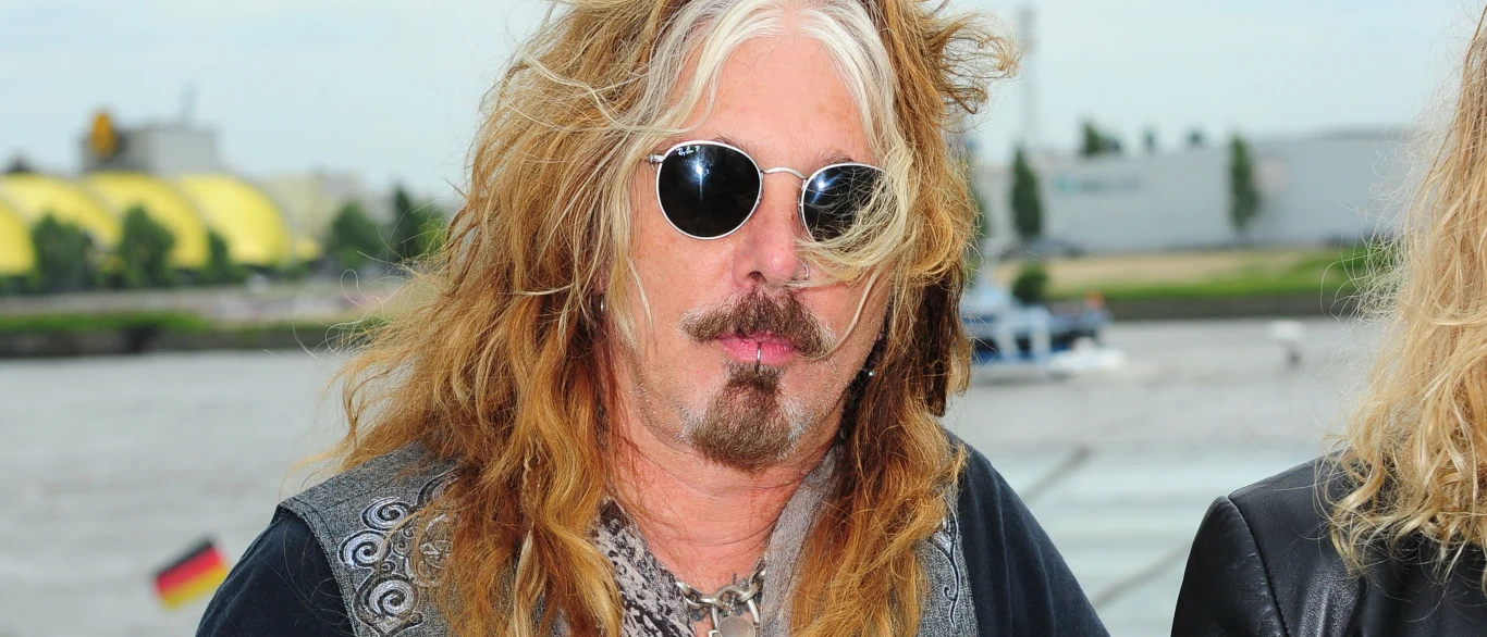 John Corabi Mężczyzna z długimi, jasnobrązowymi i lekko kręconymi włosami, okulary przeciwsłoneczne, zarost, ubrany w ciemną kurtkę, w tle rozmazany krajobraz miasta i elementy wody.