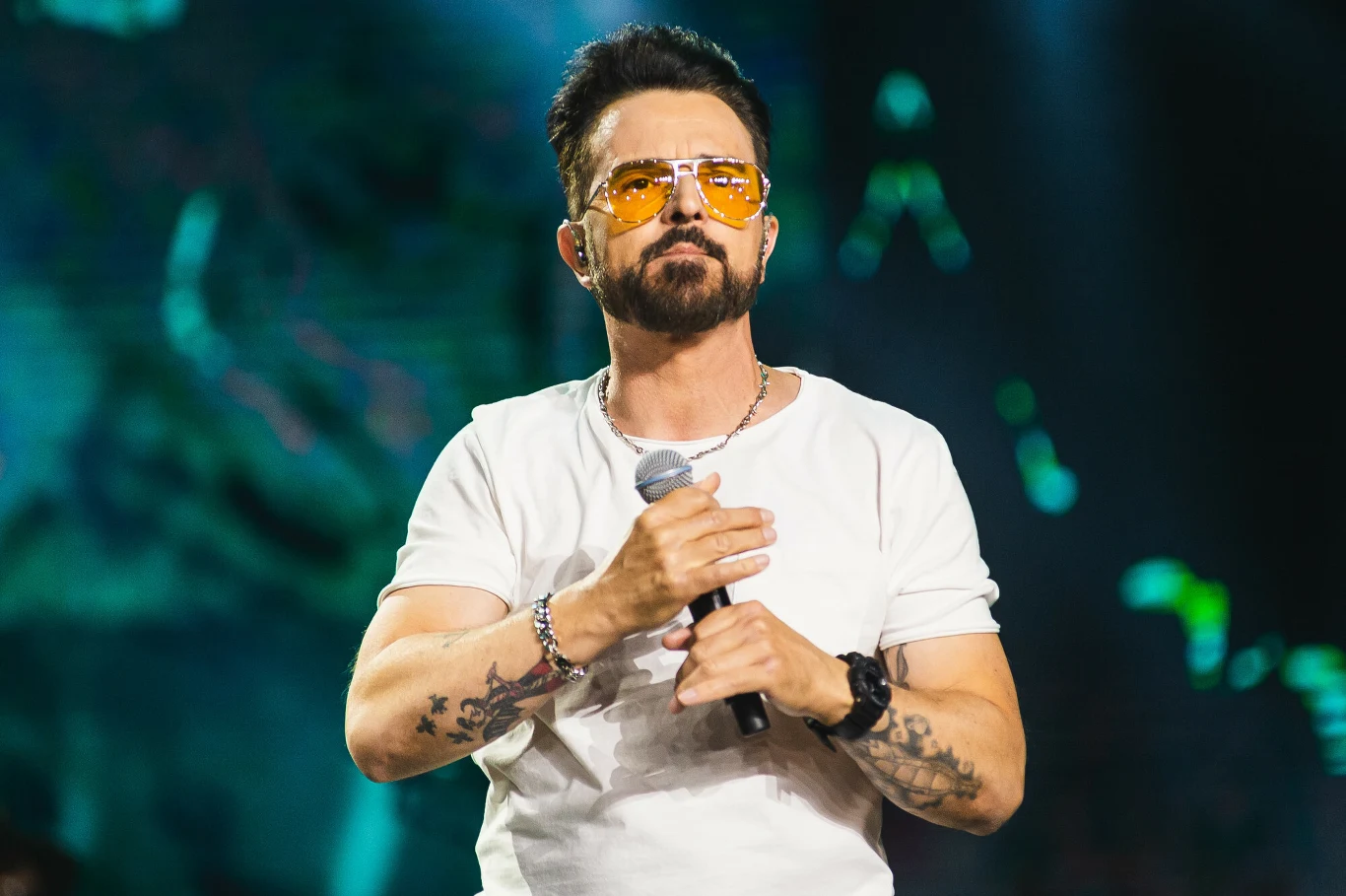 Mężczyzna z krótką brodą i tatuażami na przedramionach trzyma mikrofon, ubrany w białą koszulkę i żółte okulary przeciwsłoneczne. W tle rozmyte światła sceniczne sugerujące koncert lub występ na żywo.