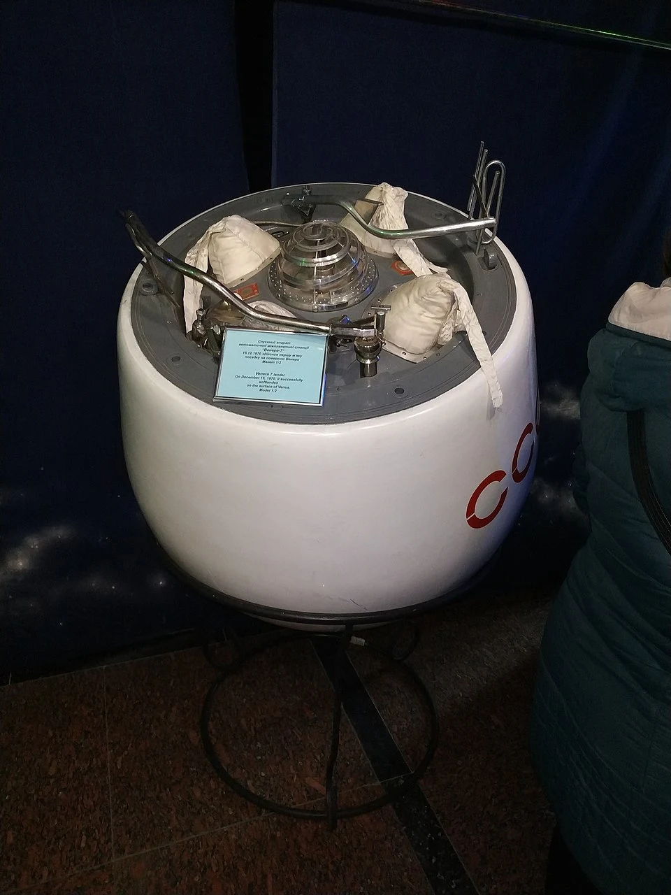 Kapsuła kosmiczna o cylindrycznym kształcie z białą zewnętrzną powłoką i widocznymi elementami technicznymi na górze, umieszczona na metalowym stojaku, częściowo zakryta przez osobę stojącą z boku.