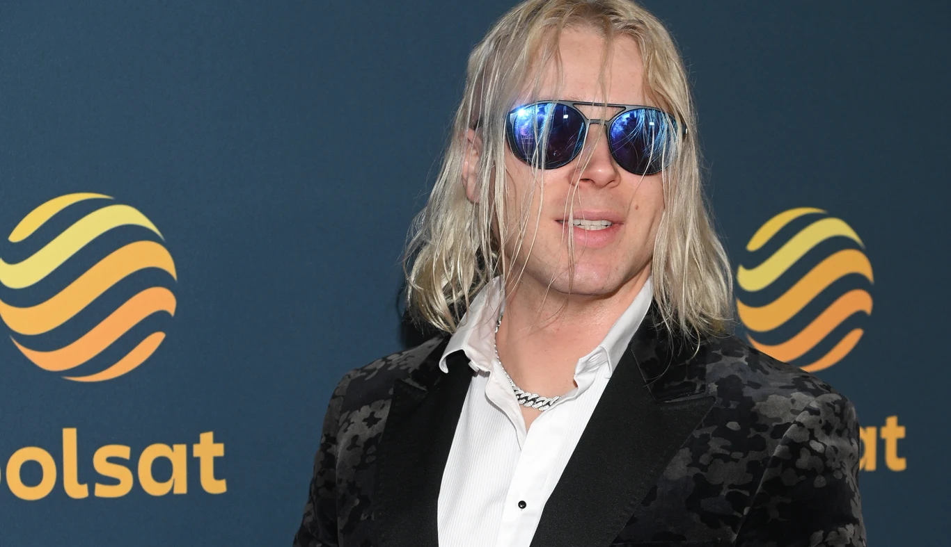 Skolim Mężczyzna w eleganckiej czarnej marynarce z białą koszulą i okularach przeciwsłonecznych stoi na tle granatowej ścianki z żółtym logotypem.