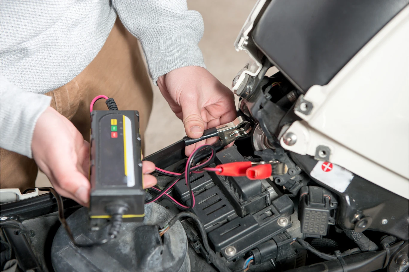 Przed pierwszym odpaleniem motocykla po zimowym przestoju warto doładować akumulator. Osoba podłącza prostownik do akumulatora w skuterze, trzymając w dłoni urządzenie ładujące oraz przewody z zaciskami.