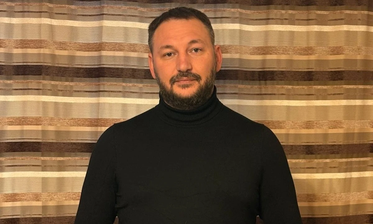 Mężczyzna o krótkich włosach i brodzie ubrany w czarny golf stoi na tle zasłony w brązowych odcieniach.