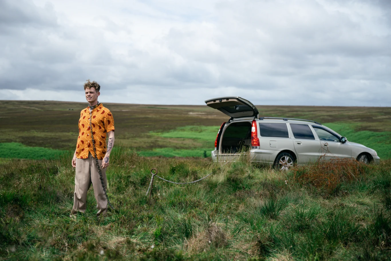 Młody mężczyzna w pomarańczowej koszuli stoi na łące obok srebrnego kombi z otwartym bagażnikiem, w tle rozległy krajobraz polany pod chmurami.