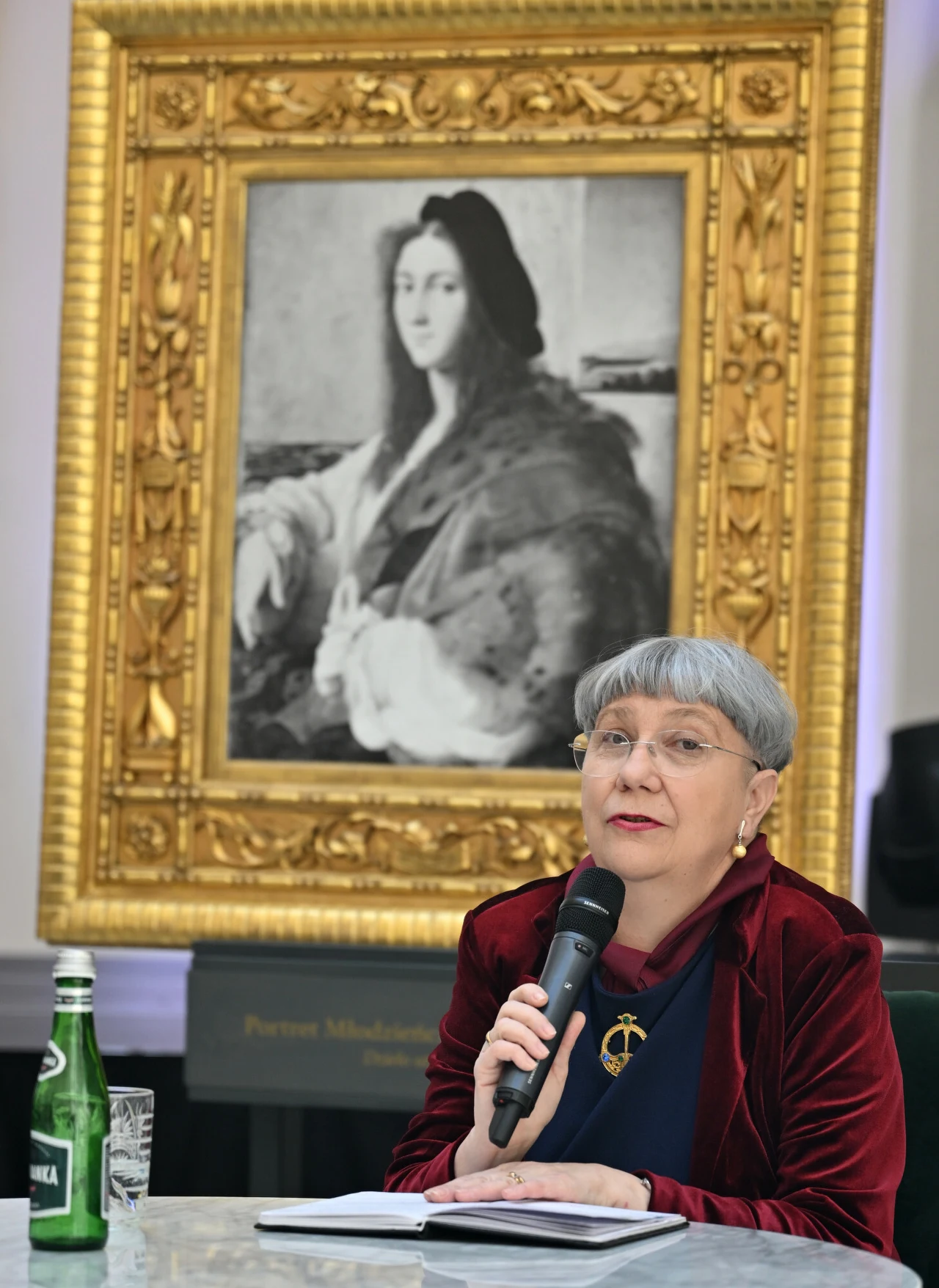 Katarzyna Płonka-Bałus: "W przypadku "Portretu młodzieńca” mamy do czynienia ze stratą wojenną - dziełem zaginionym. Nie wiemy czy bezpowrotnie" Starsza kobieta z mikrofonem w ręku siedzi przy stole, na którym stoi zielona butelka i szklanka wody. W tle wisi duży, ozdobny portret kobiety w złotej ramie.