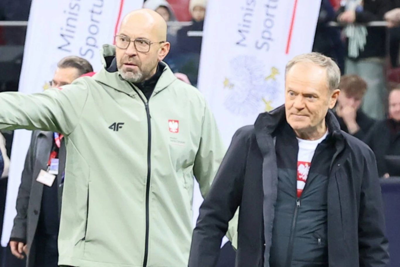 Minister sportu, Jakub Rutnicki i premier Donald Tusk Dwóch mężczyzn w ubraniach sportowych podczas wydarzenia sportowego, jeden wskazuje coś ręką, w tle banery z napisem Ministerstwo Sportu.