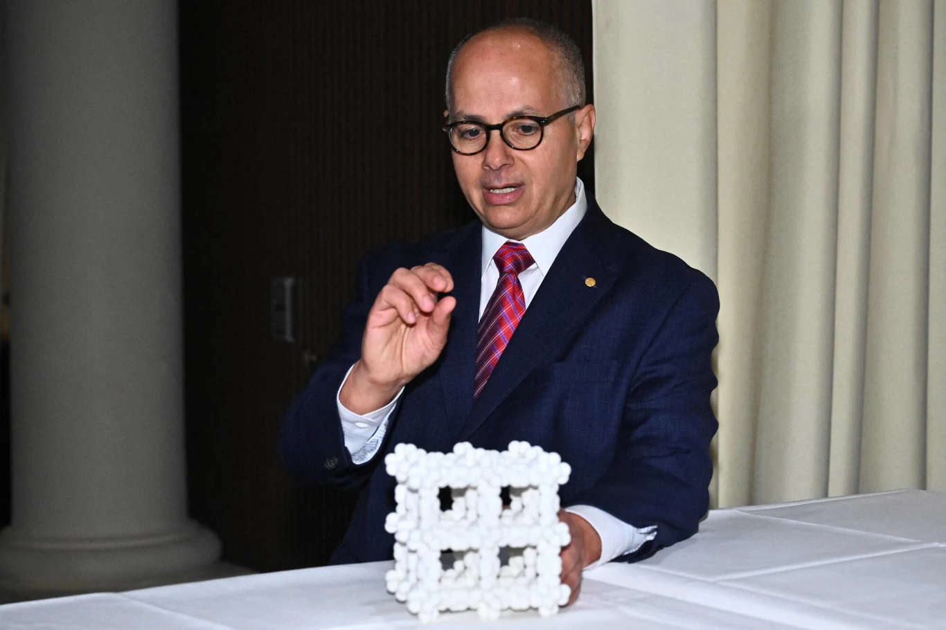 Noblista opracował maszynę, która wyciąga wodę z powietrza. Przełom w walce z globalnym kryzysem wodnym? Elegancko ubrany mężczyzna w garniturze i okularach trzyma przestrzenną strukturę wykonaną z białych kulek i prętów, siedząc przy białym stole na tle zasłon.