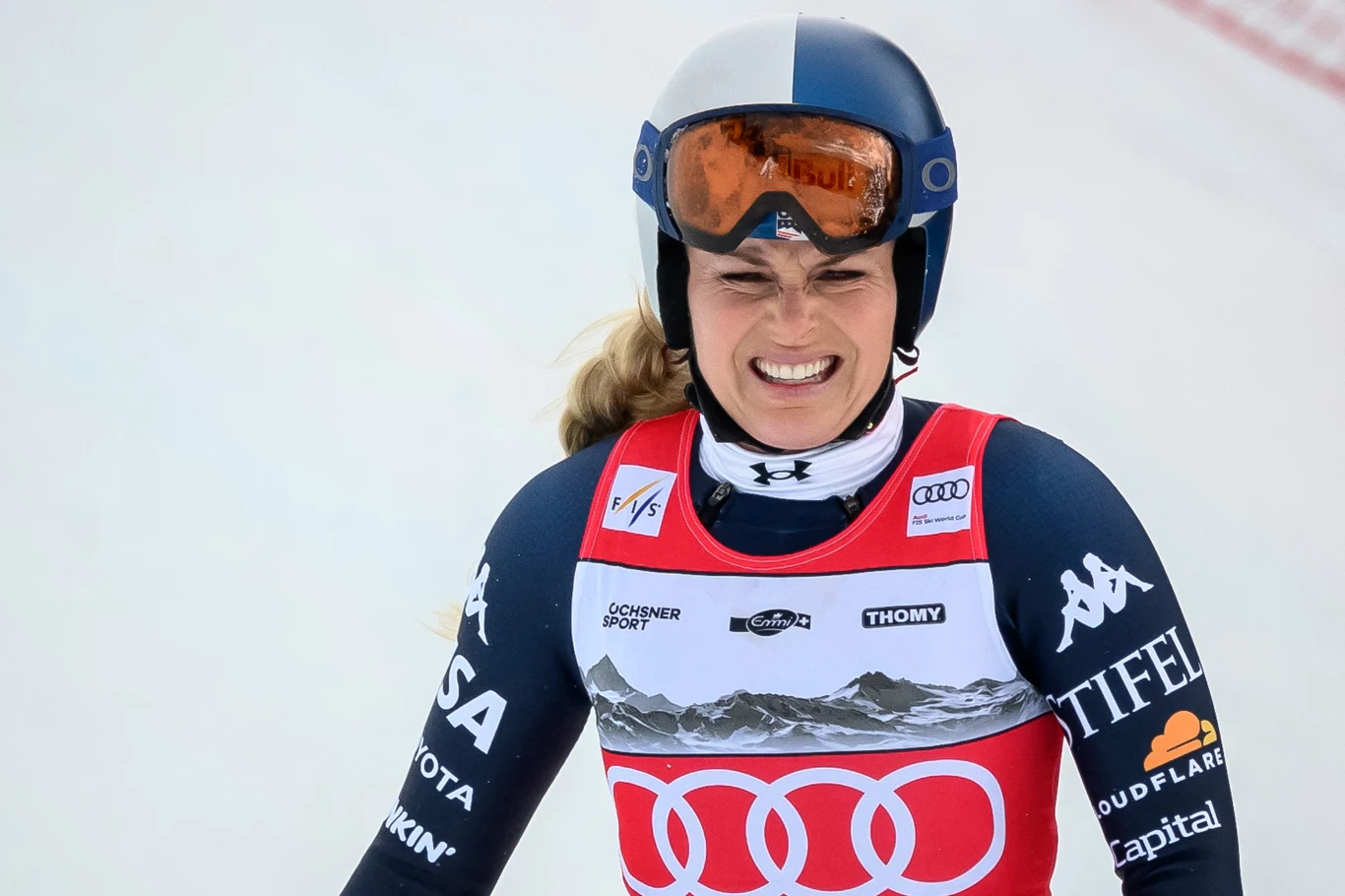 Lindsey Vonn Zawodniczka w stroju narciarskim i kasku ochronnym z goglami na czole stoi na stoku, ma na sobie numer startowy i sportową odzież z logotypami sponsorów, uśmiecha się energicznie po zakończonym wyścigu.