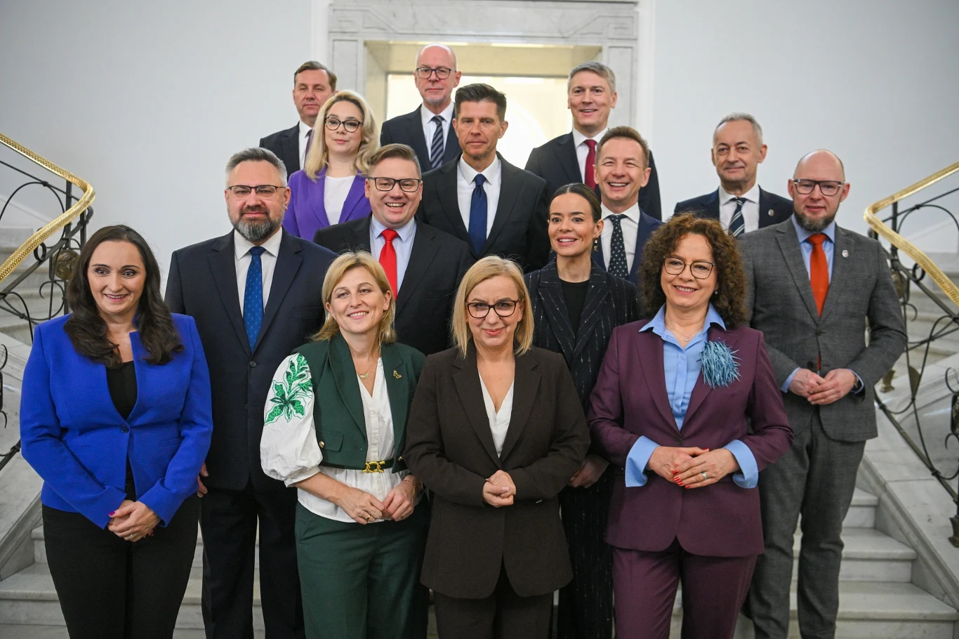 Grupa elegancko ubranych osób ustawionych na klatce schodowej w reprezentacyjnym wnętrzu gmachu, wszyscy patrzą w stronę obiektywu i uśmiechają się, atmosfera formalna i oficjalna.