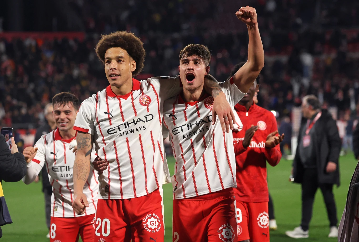 Grupa piłkarzy drużyny piłkarskiej Girona FC w biało-czerwonych pasiastych koszulkach świętuje zwycięstwo na murawie stadionu, jeden z zawodników unosi triumfalnie rękę, a wokół widać innych członków zespołu oraz kibiców na trybunach.