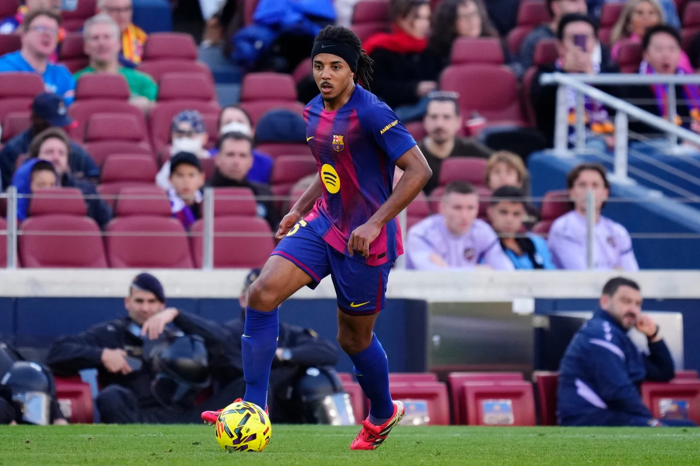 Piłkarz w stroju FC Barcelony prowadzi piłkę na boisku podczas meczu, w tle widoczna pełna trybuna kibiców.