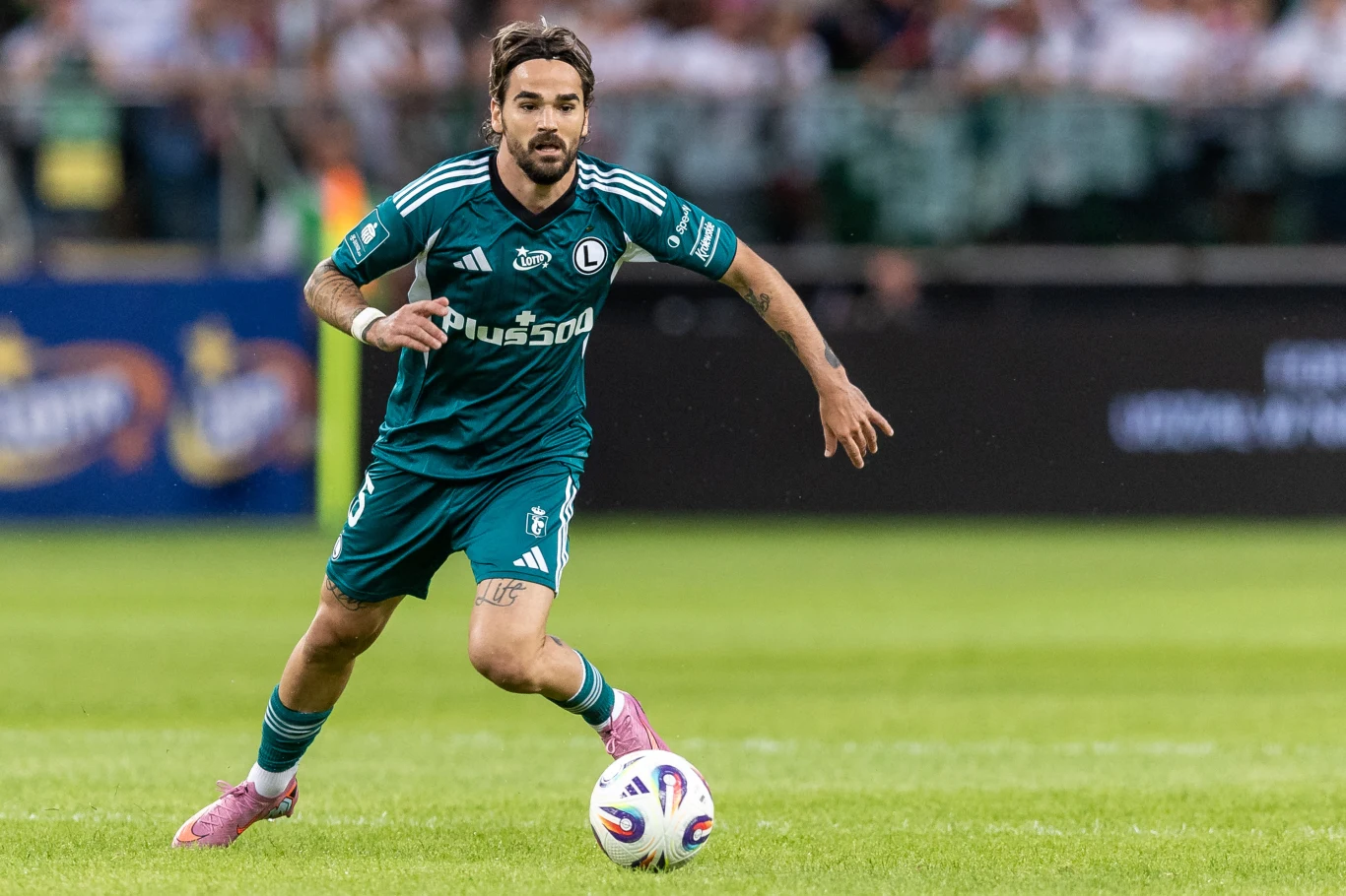 Piłkarz w zielonym stroju klubowym biegnie po boisku z piłką u swoich stóp podczas meczu piłkarskiego, w tle rozmyty stadion z kibicami.