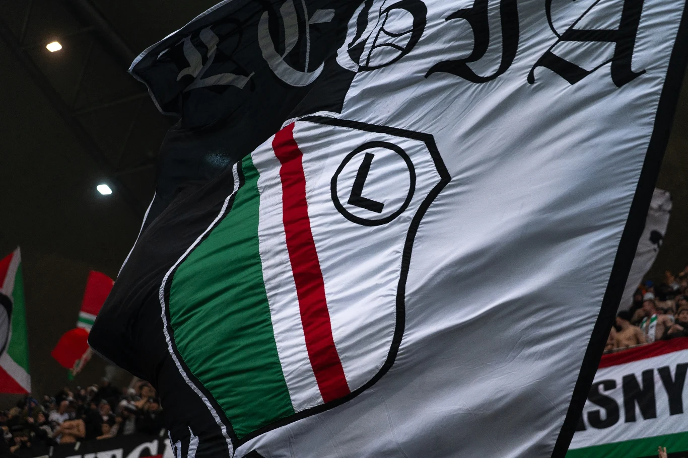 Duża flaga z herbem klubu piłkarskiego Legia Warszawa w biało-zielono-czerwono-czarnych barwach powiewająca na stadionie, w tle widoczni kibice i inne flagi.