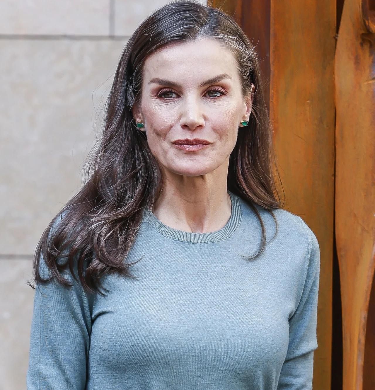 Królowa Letizia wie, w jak delikatny sposób zwrócić na siebie uwagę Kobieta z długimi brązowymi włosami ubrana w jasnozielony sweter stoi na tle jasnej ściany oraz brązowej, drewnianej lub metalowej dekoracji.