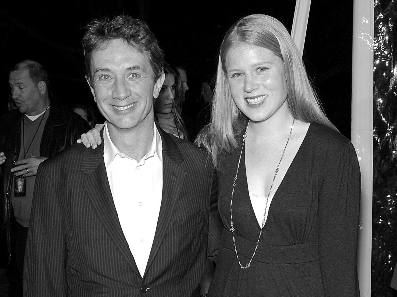 Martin Short ze swoją córką Katherine Hartley Short Mężczyzna w garniturze i młoda kobieta w eleganckiej sukience pozują razem do zdjęcia, oboje uśmiechnięci, w tle kilka osób w mało wyraźnym otoczeniu.