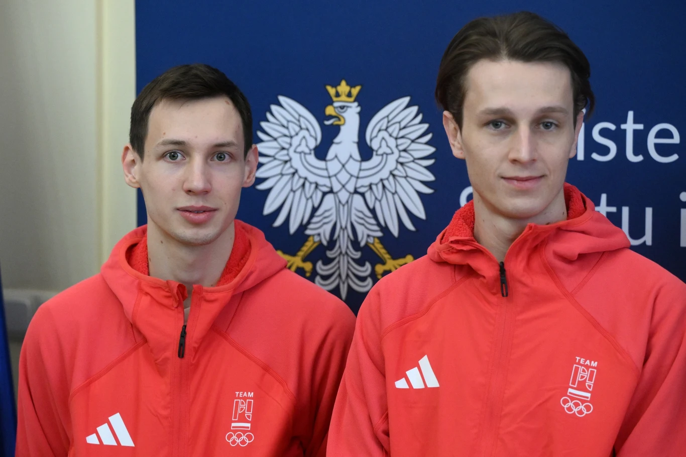 Dwóch młodych mężczyzn w czerwonych sportowych bluzach z logiem polskiej reprezentacji olimpijskiej, stojących na tle niebieskiego banera z białym orłem.