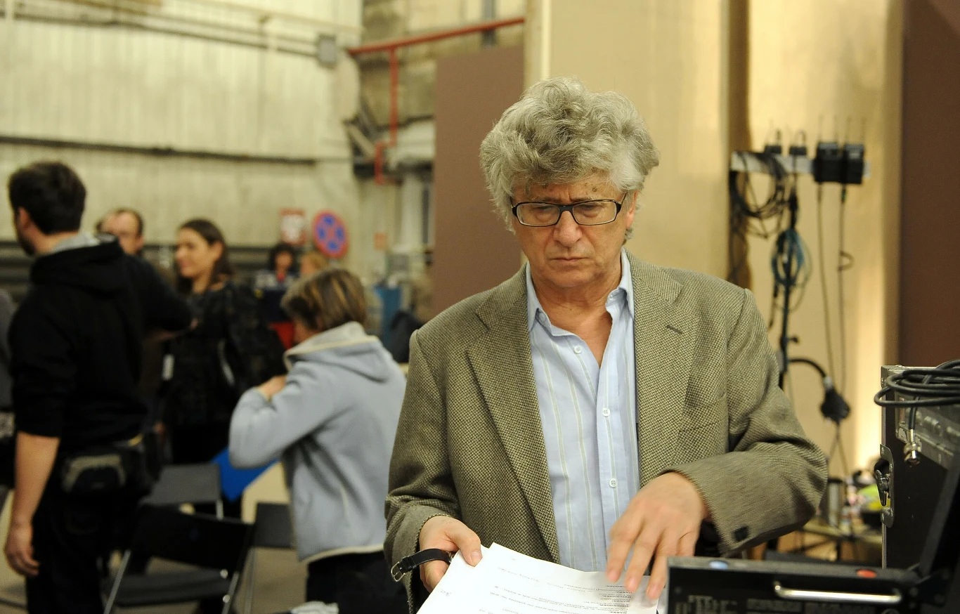 Mężczyzna w okularach i marynarce stoi przy biurku z dokumentami, w tle kilka osób w rozmowie, industrialne wnętrze i sprzęt techniczny.