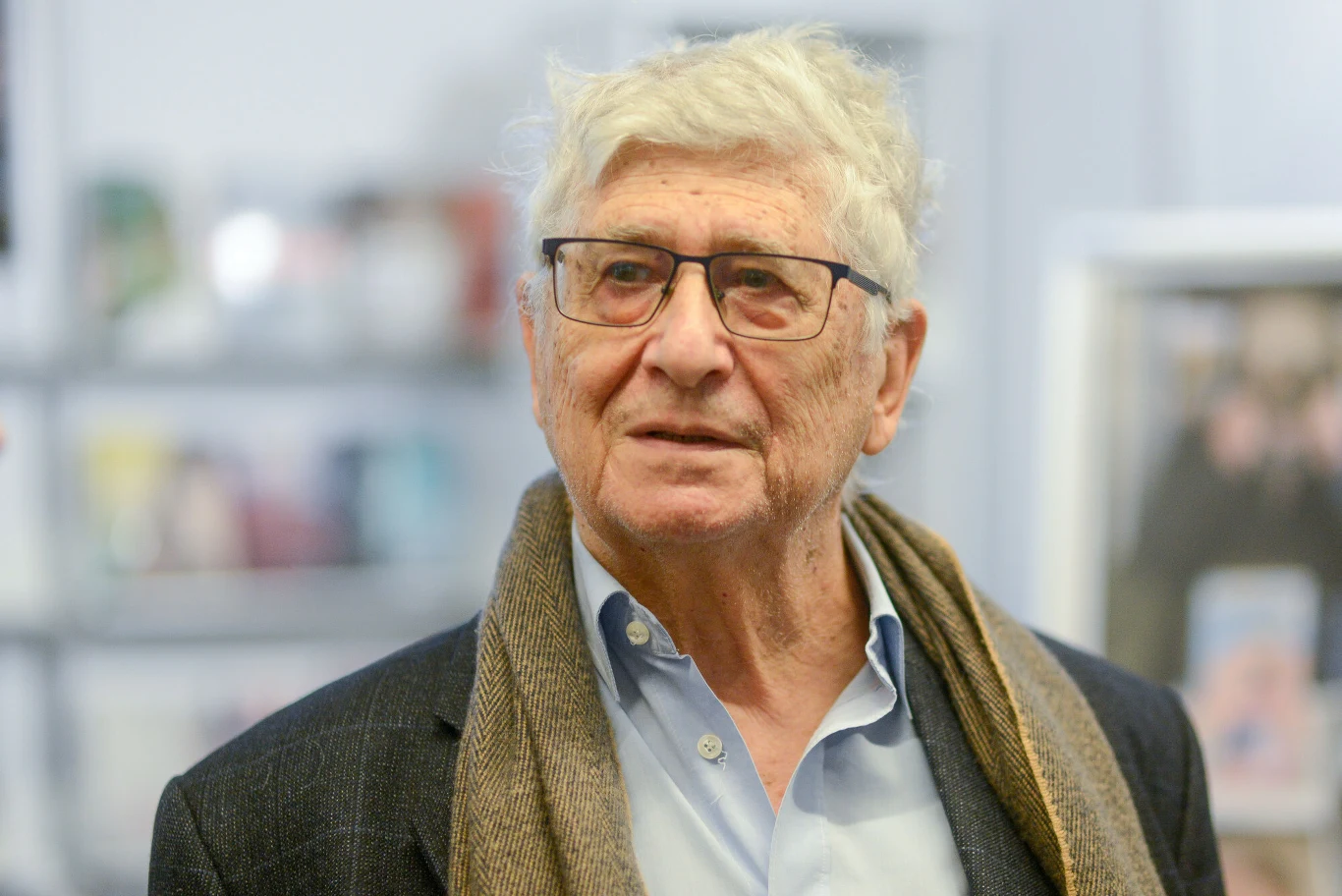 Starszy mężczyzna w okularach oraz szaliku stoi w dobrze oświetlonym, prawdopodobnie publicznym miejscu, na jego twarzy widoczny jest spokojny wyraz.