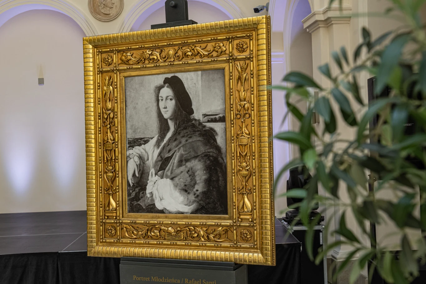 Czy obraz uda się odnaleźć? Złocona, ozdobna rama z czarno-białym portretem kobiety w renesansowym stroju, ustawiona na sztaludze w jasno oświetlonym pomieszczeniu, w tle fragment sceny i roślinność.