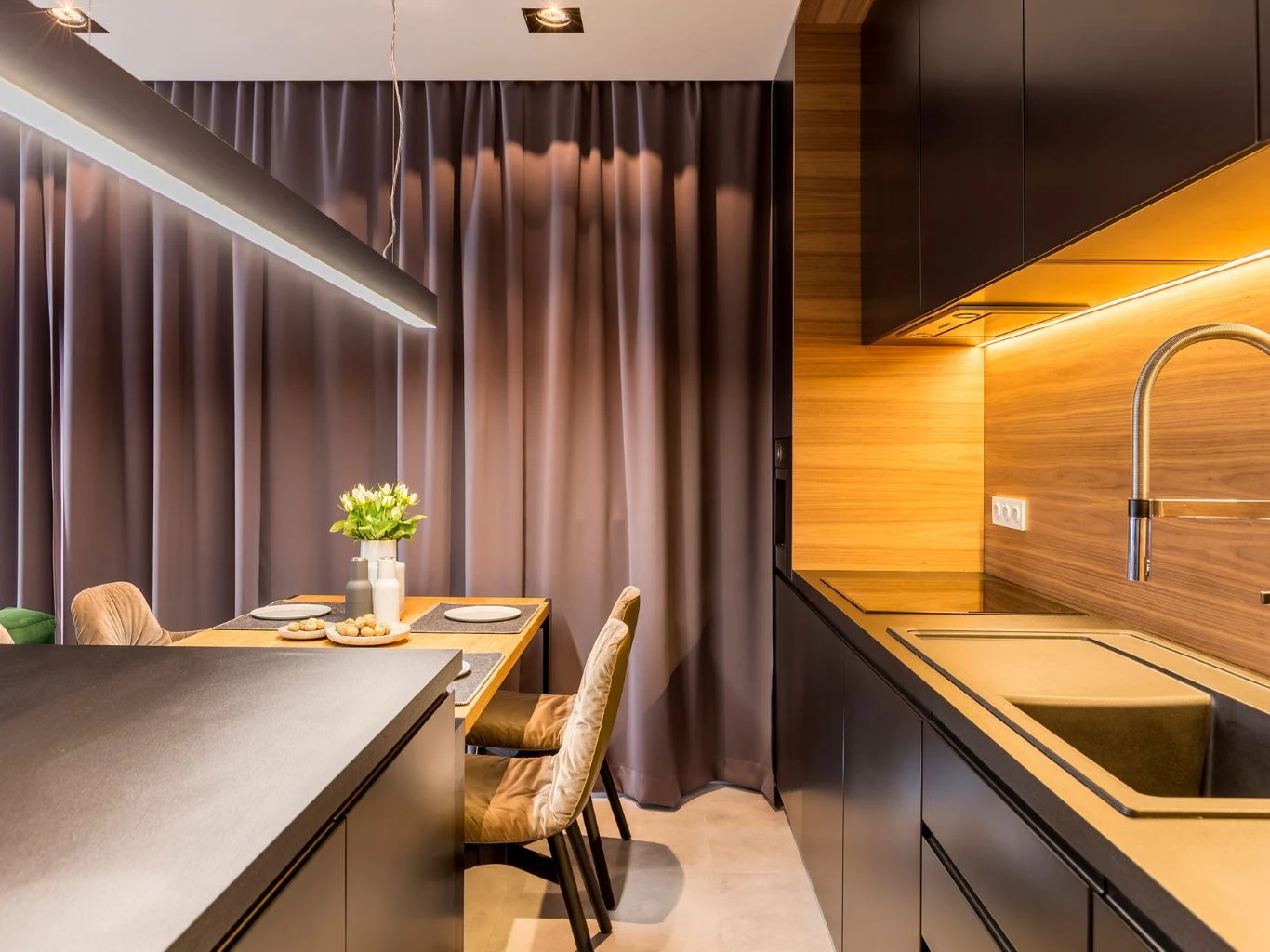 Technologia ta jest uwielbiana przez dekoratorów wnętrz i architektów oświetleniowych, ponieważ daje niemal nieograniczone możliwości aranżacyjne. Nowoczesna kuchnia z czarnymi szafkami, drewnianymi elementami i zlewem pod zabudową. Przy jadalnianym stole ustawiono cztery krzesła, na stole znajduje się biały wazon z kwiatami oraz zastawa stołowa. Tło stanowią ciemne zasłony.