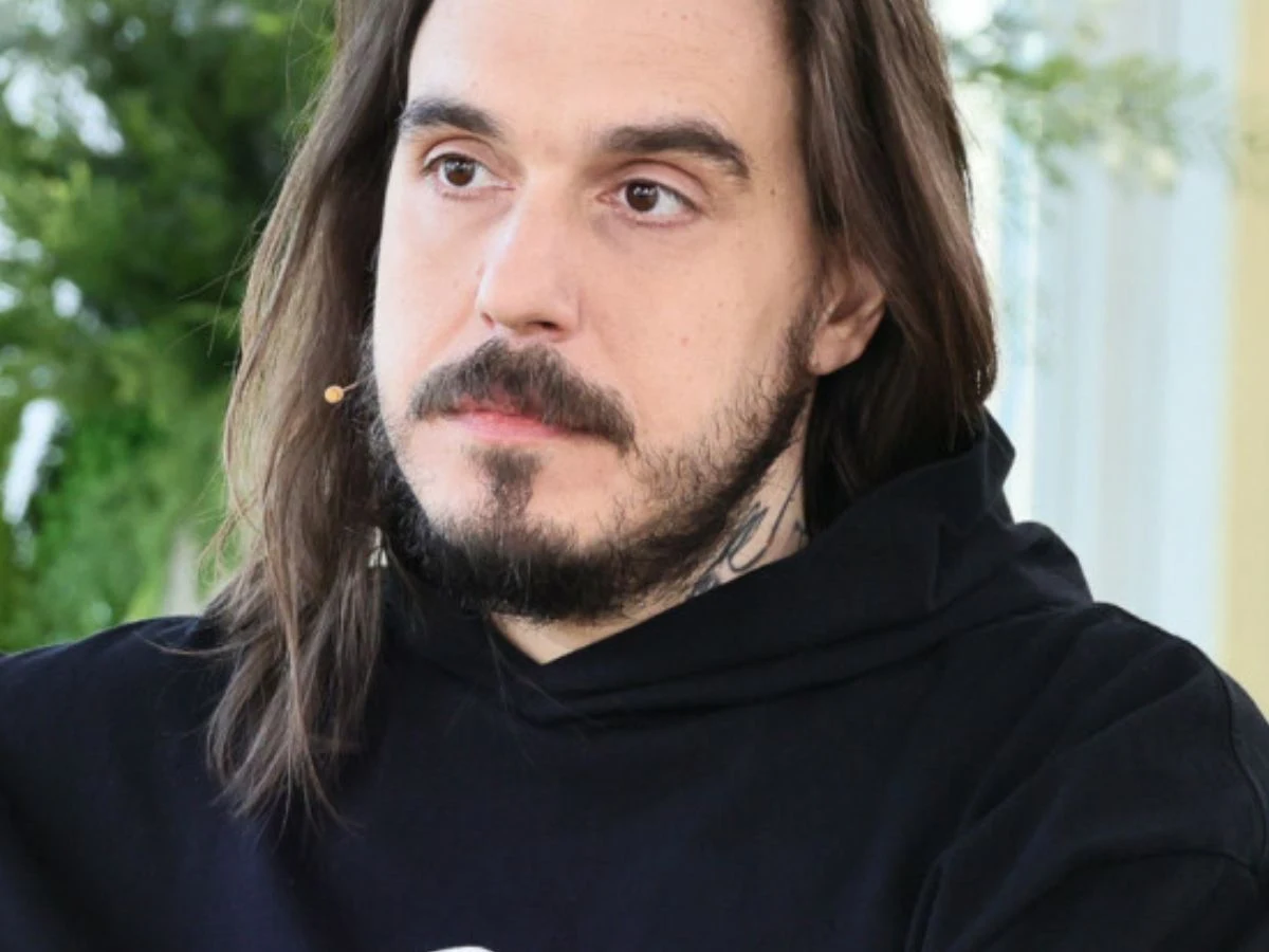 Miuosh Mężczyzna z długimi włosami i brodą, ubrany w czarną bluzę z kapturem, z mikrofonem na policzku, na tle roślinności wewnętrznej.