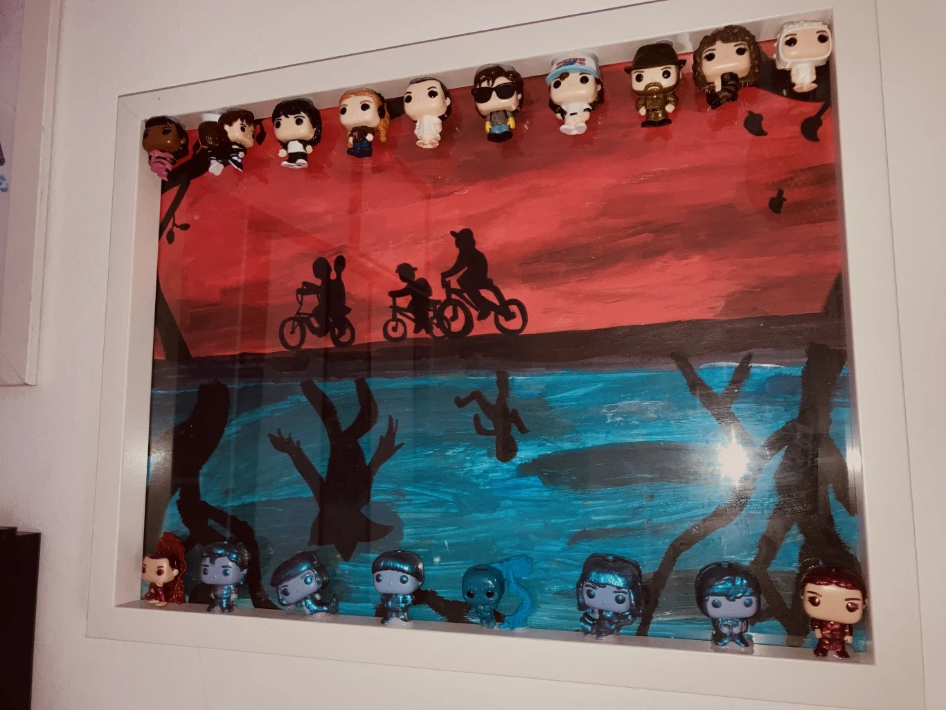 Kolekcja figurek przedstawiających bohaterów umieszczona w białej gablocie na tle malowanego obrazu z sylwetkami dzieci jadących na rowerach przez las przy czerwono-niebieskim niebie.