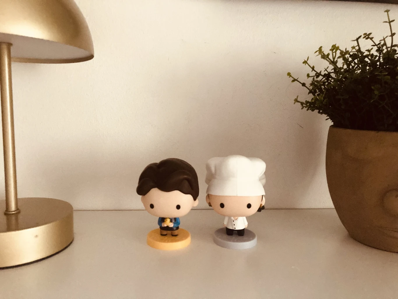 Dwie figurki w stylu chibi stoją na białej powierzchni; jedna z postaci ubrana jest w niebieski strój, druga w strój kucharza z białą czapką. Po bokach widać fragment lampki oraz doniczkę z zieloną rośliną.