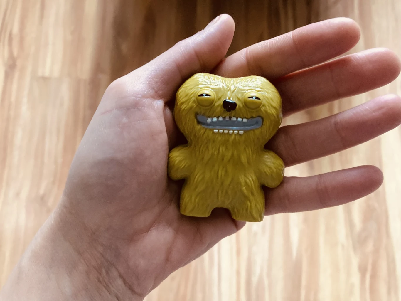 Żółta, futrzasta figurka o uśmiechniętej, karykaturalnej twarzy i dużych oczach leży na otwartej dłoni z jasnym drewnianym tłem w tle.