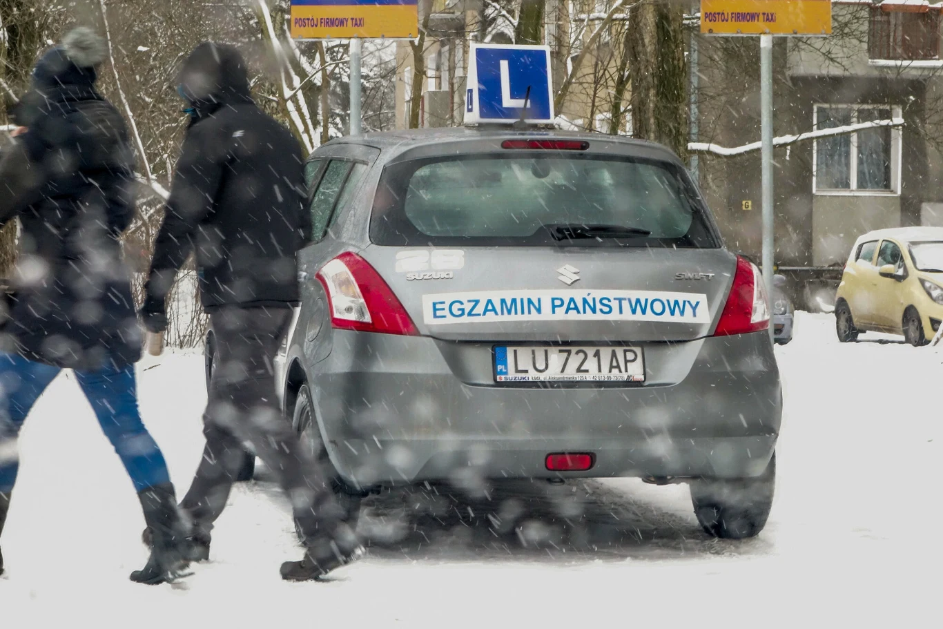 Samochód egzaminacyjny podczas części praktycznej. Spór o użycie lewego kierunkowskazu trafił aż do sądu. Samochód egzaminacyjny do nauki jazdy, oznaczony literą L i napisem 'Egzamin Państwowy', zaparkowany na zaśnieżonej ulicy, obok przechodzą dwie osoby podczas intensywnych opadów śniegu.