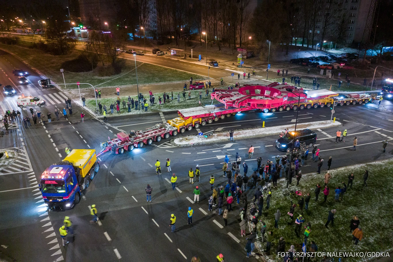 Przewozy nienormatywne obejmują ładunki, których masa, długość lub szerokość przekraczają dopuszczalne normy dla standardowych pojazdów. Nocny transport ogromnej konstrukcji przez skrzyżowanie w mieście, otoczony liczną grupą obserwatorów oraz zabezpieczany przez pojazdy eskortujące i służby porządkowe. Oświetlenie uliczne podkreśla skalę operacji oraz obecność ciekawskich mieszkańców.