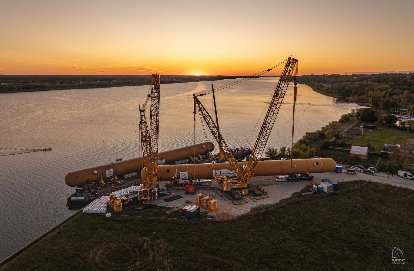 W ostatnich latach po polskich drogach poruszały się zestawy, których masa dorównuje dużym jednostkom pływającym czy samolotom transportowym. Wielkogabarytowe dźwigi budowlane ustawione nad brzegiem rzeki unoszą i pozycjonują ogromne, cylindryczne elementy konstrukcyjne. W tle spokojna powierzchnia wody oraz rozległe tereny zielone oświetlane zachodzącym słońcem.