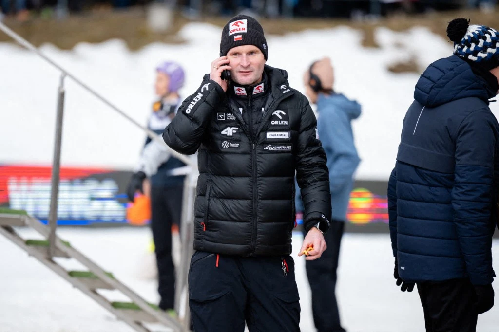 Jaka przyszłość czeka Macieja Maciusiaka? Mężczyzna w czarnej kurtce sportowej z logotypami sponsorów stoi na śniegu, rozmawia przez telefon i trzyma w ręku mały przedmiot, w tle osoby oraz elementy infrastruktury zimowej imprezy sportowej.