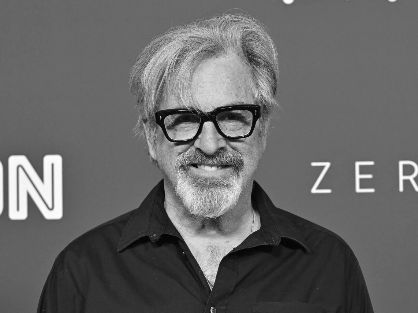 Robert Carradine Starszy mężczyzna z siwymi włosami i brodą, uśmiechnięty, w okularach o grubych oprawkach, ubrany w ciemną koszulę, stoi na tle z rozmytymi napisami.