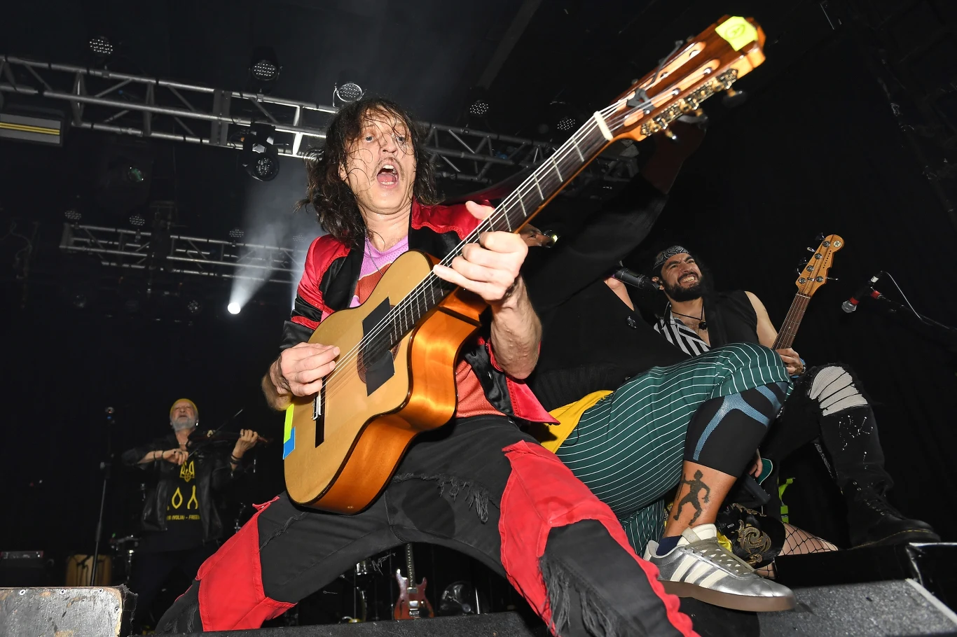 Gogol Bordello na żywo to istny ogień Energetyczny gitarzysta w kolorowym stroju gra na scenie, otoczony przez innych muzyków, dynamiczne światła koncertowe podkreślają atmosferę występu, a publiczność rozświetlona reflektorami podziwia ich występ.