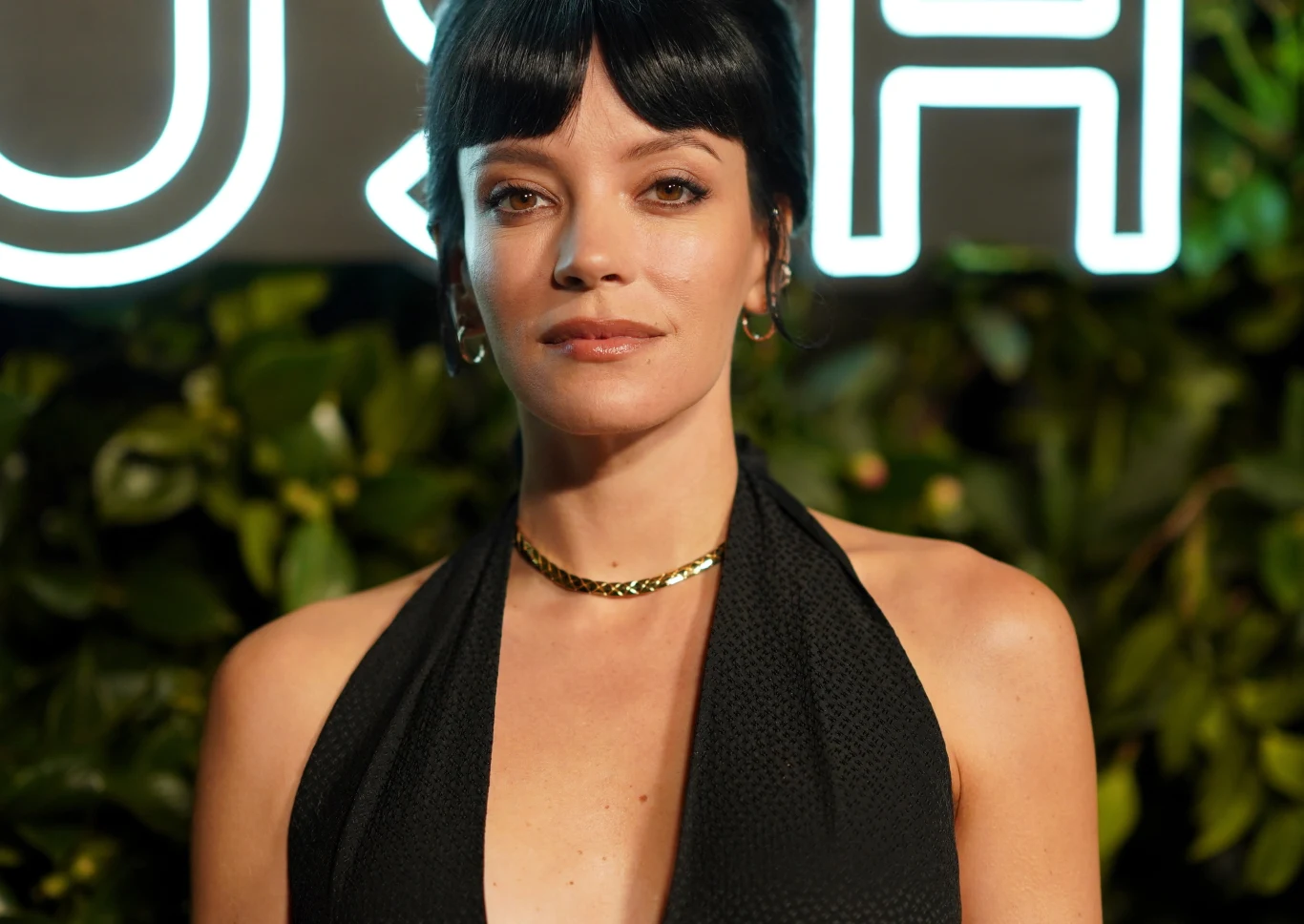 Lily Allen Kobieta z krótkimi, ciemnymi włosami, ubrana w elegancką, czarną suknię z głębokim dekoltem oraz złoty łańcuszek, stoi na tle zieleni i jasnego napisu.