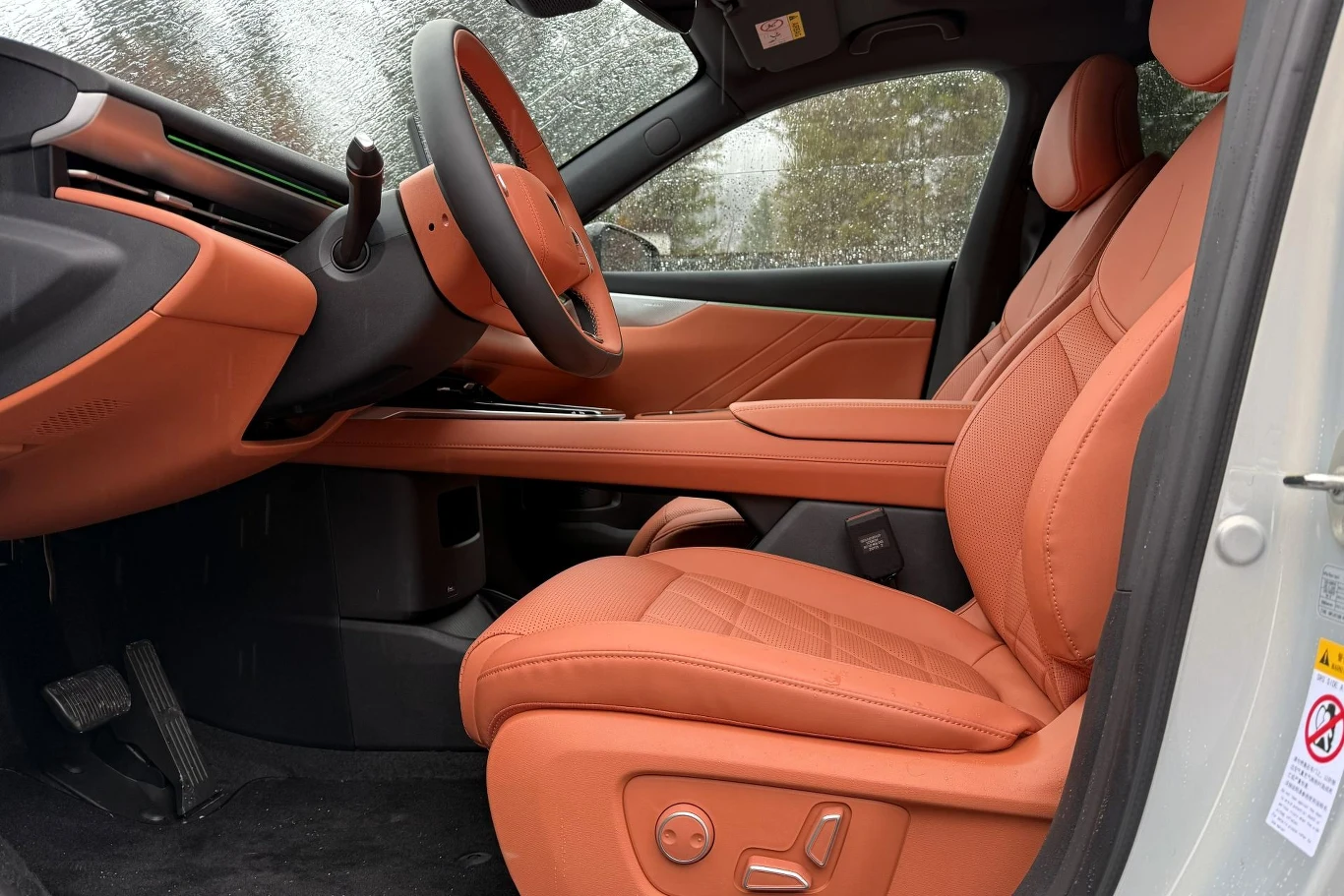Przednie fotele już w standardzie wyposażone są w elektryczną regulację. Luksusowe wnętrze samochodu z pomarańczową skórzaną tapicerką, widoczne przednie siedzenie, deska rozdzielcza oraz kierownica, na zewnątrz pada deszcz.