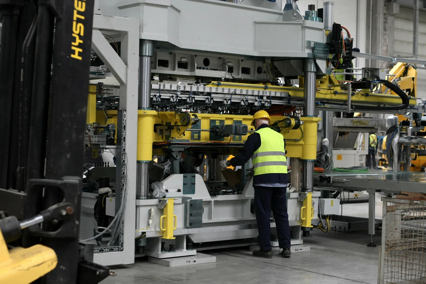 Wcześniejsze emerytury i dobrowolne odprawy. Gigant zamyka zakład | Fot. ilustracyjna Pracownik w kamizelce odblaskowej i kasku obsługuje dużą, przemysłową maszynę w hali produkcyjnej otoczonej innymi urządzeniami oraz elementami technicznymi.