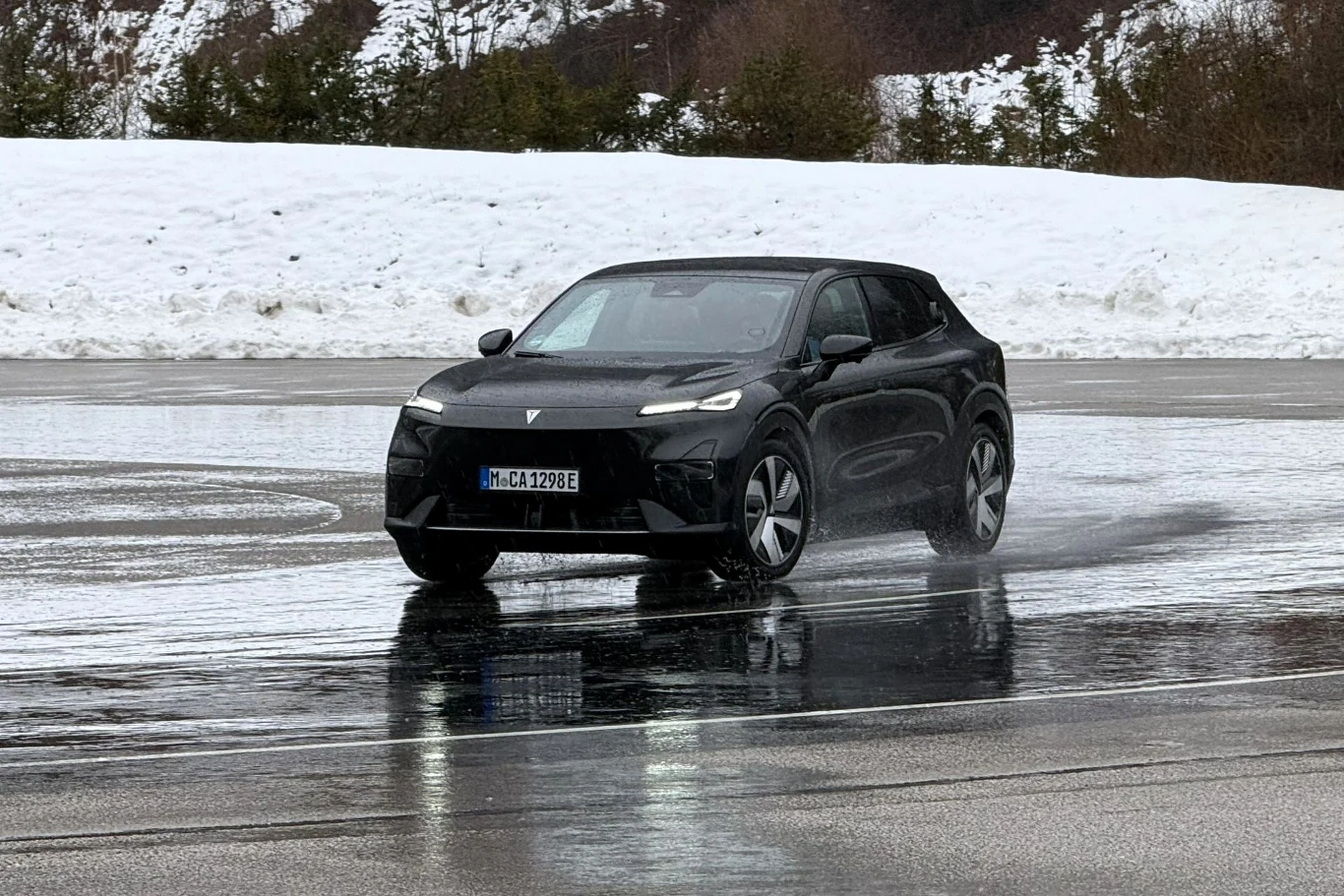 W kontrolowanych warunkach testowych wyposażony w napęd na tylne koła Changan Deepal S05 nie zawsze radził sobie ze śliską nawierzchnią. Czarny samochód elektryczny jedzie po mokrym asfalcie, wokół znajduje się śnieg oraz niskie drzewa i krzewy. Auto odbija się od nawierzchni pokrytej wodą, w tle widoczne są wzgórza.